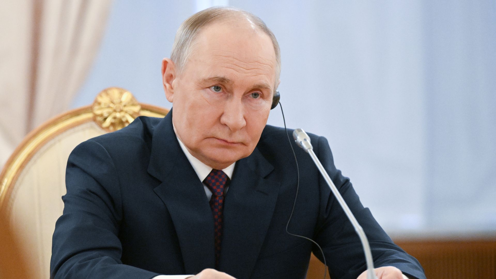 A man in a dark suit and patterned tie sits at a desk with a microphone and papers, wearing an earpiece, against a light blue background with gold and beige curtains.
