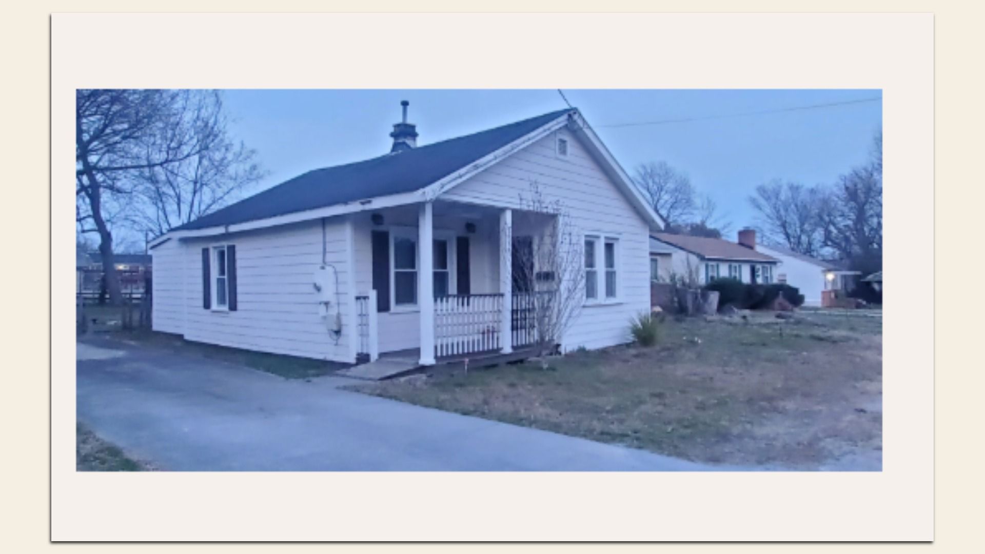 White, single-story ranch house with a small covered front porch supported by posts. Dark pitched roof with a chimney, leafless trees in the yard, a driveway, and neighboring homes under a blue sky.