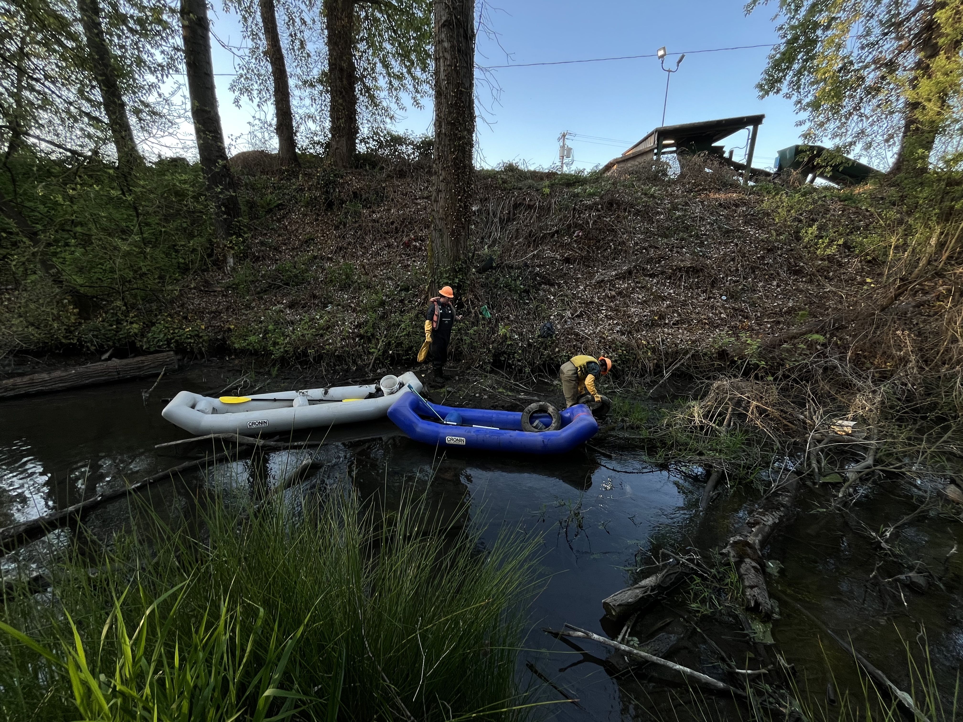 Two workers in orange helmets stand by a river with white and blue inflatable boats; a leaf-covered slope rises behind them with trees and a wooden shed on a hill, under a clear blue sky.