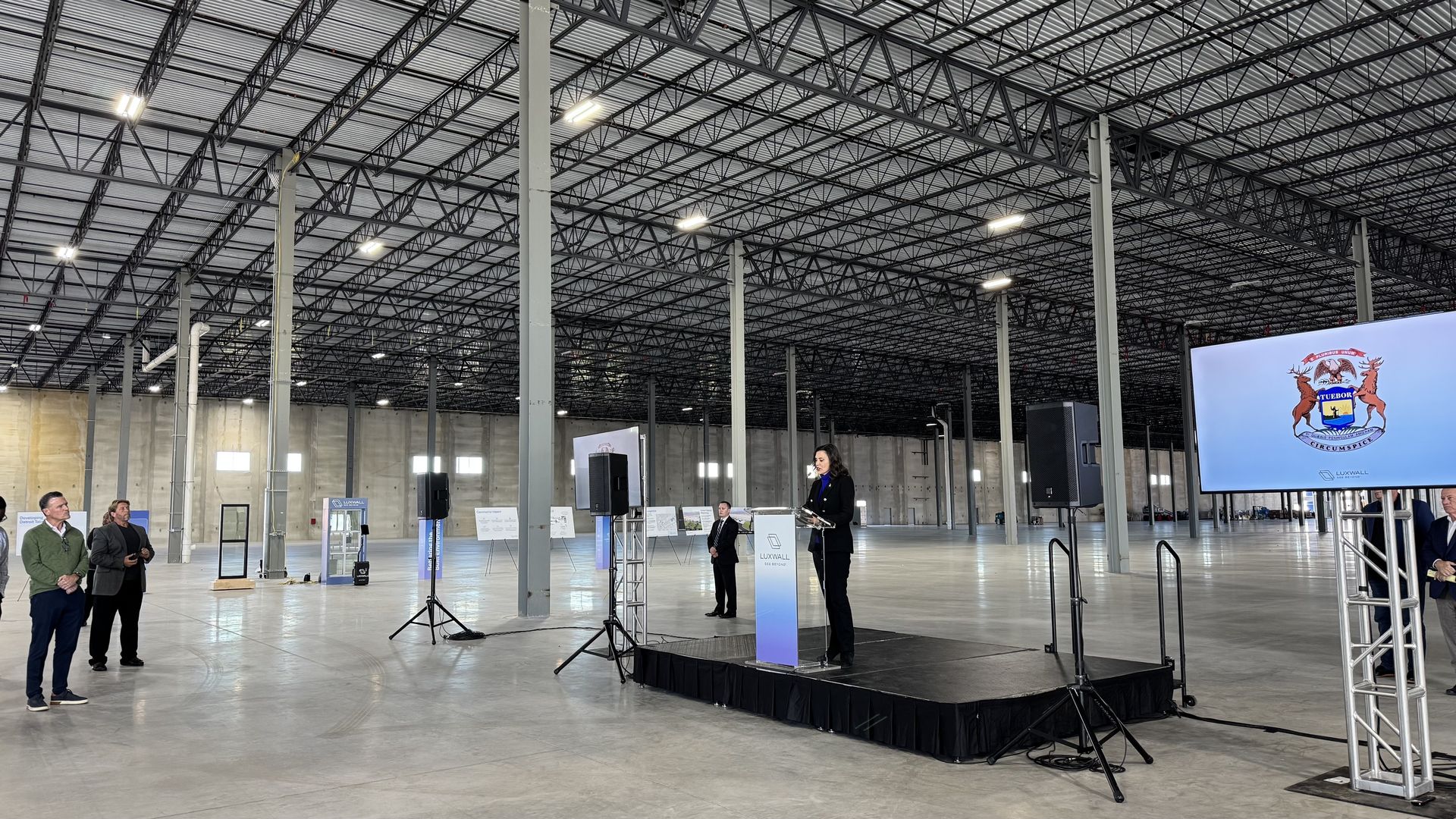 Gov. Whitmer is shown speaking at a podium surrounded by a giant empty plant building