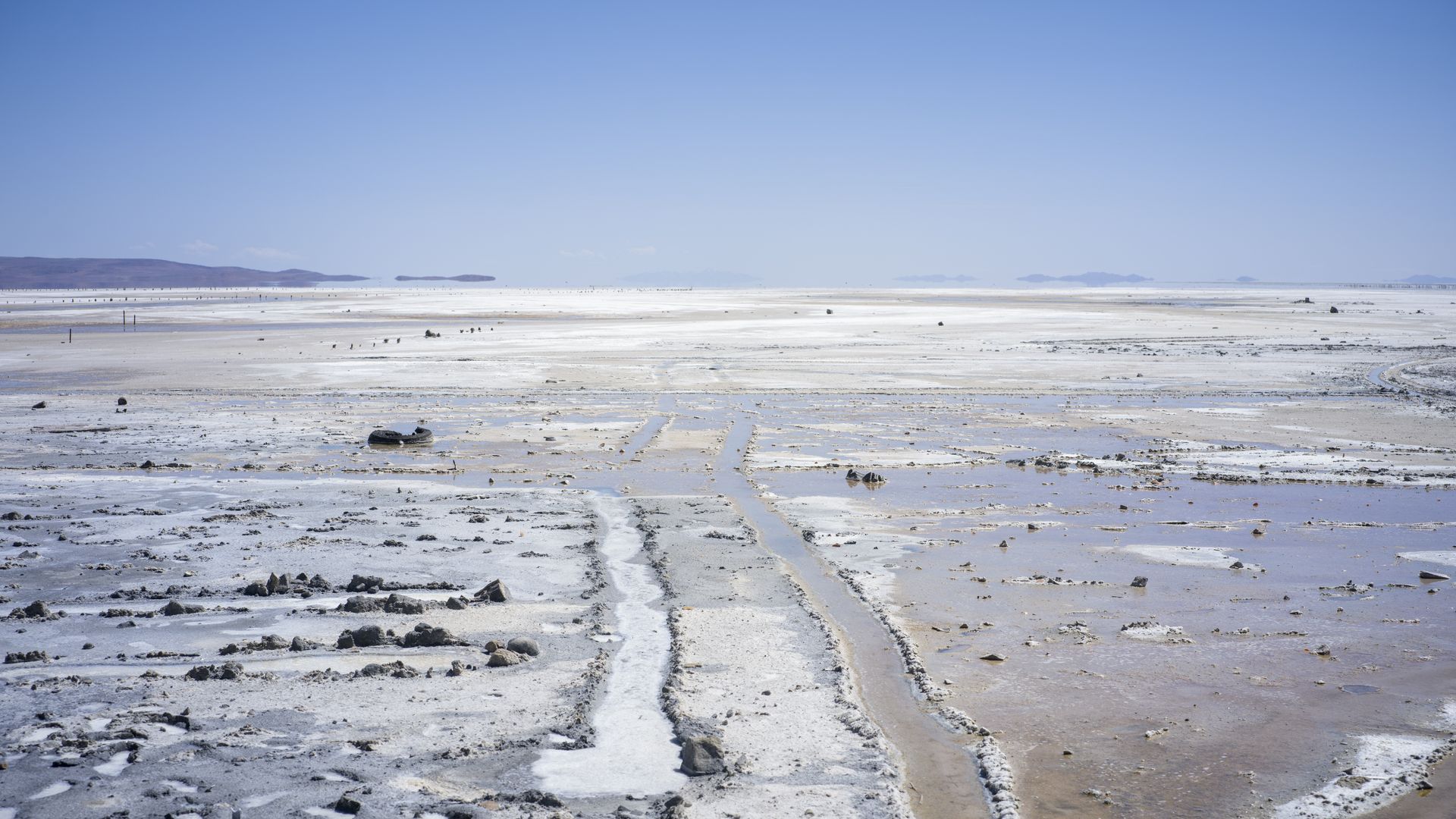 Expansive salt flat with white crust, shallow water channels, scattered rocks, and distant blue mountains beneath a clear blue sky.