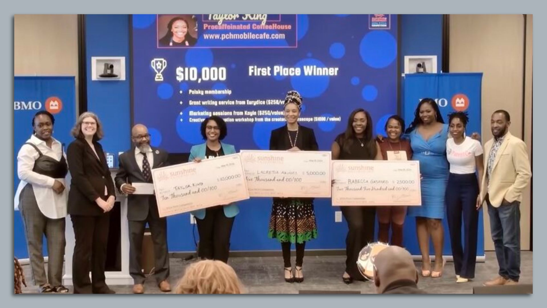 Ten people on stage, three in the middle holding large checks, standing in front of a blue screen.