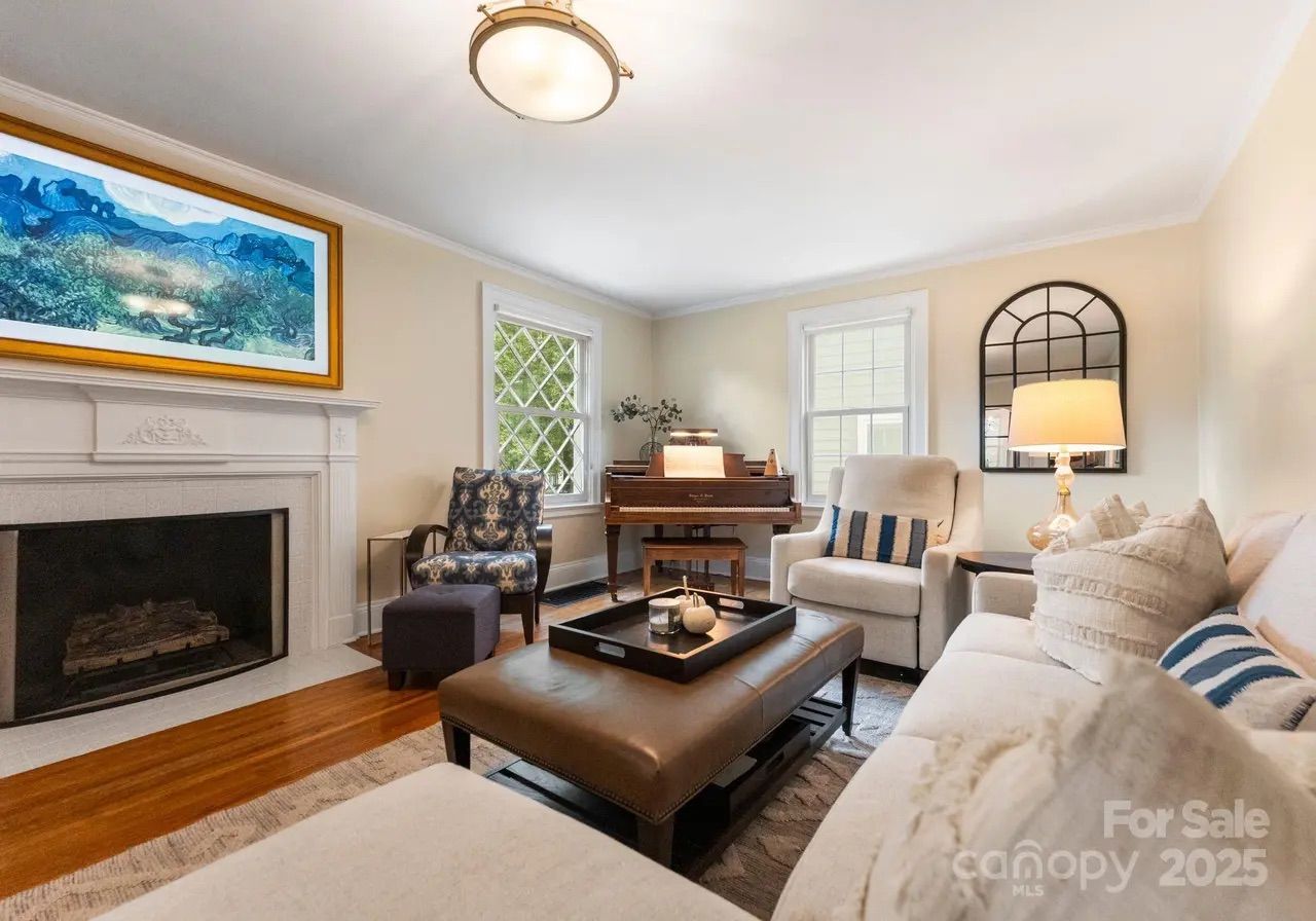 Cozy living room with beige walls, a white fireplace, blue patterned armchair, cream sofa with striped pillows, brown leather ottoman, piano, and a lit table lamp under a window mirror.