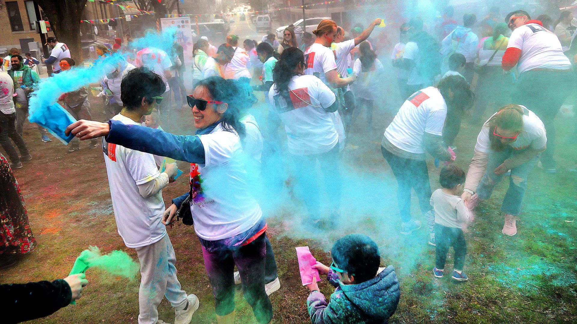 Massive outdoor color festival on a city street: participants in white shirts throw blue and green powder, smoke fills the air as kids and adults celebrate amid bunting and buildings.