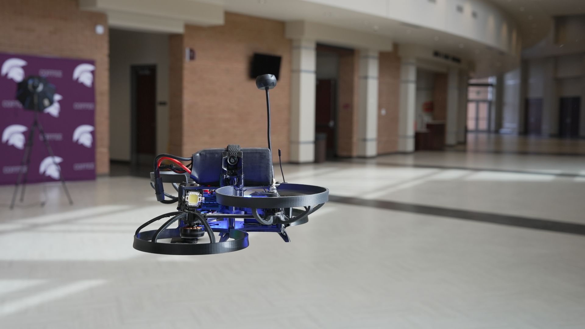 Blue circular hovering robot with a vertical mast and small round camera on top, indoors in a spacious atrium with brick walls and a purple event backdrop.