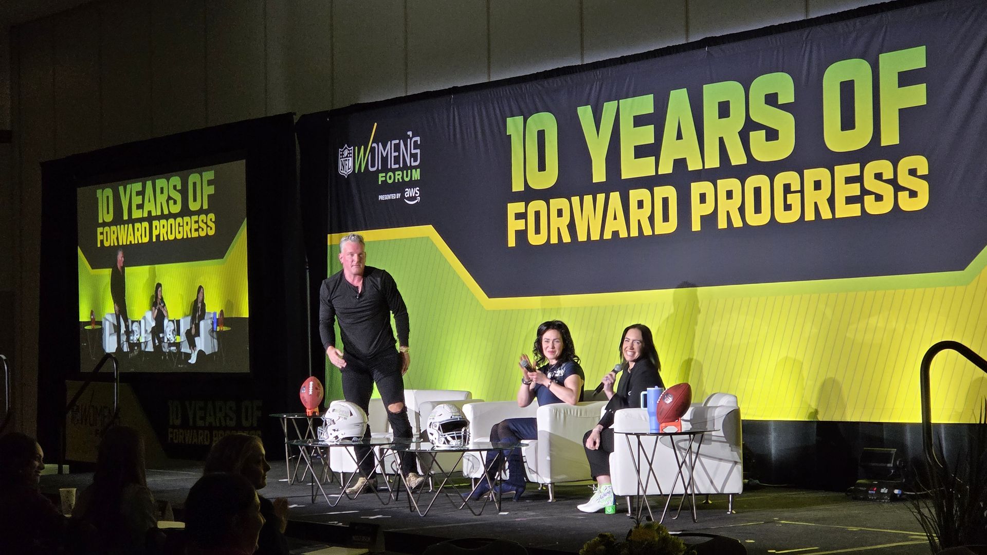 A man and two women on stage at the NFL Women's Forum with a large backdrop reading 10 Years of Forward Progress in green and yellow, football helmets and balls on tables.