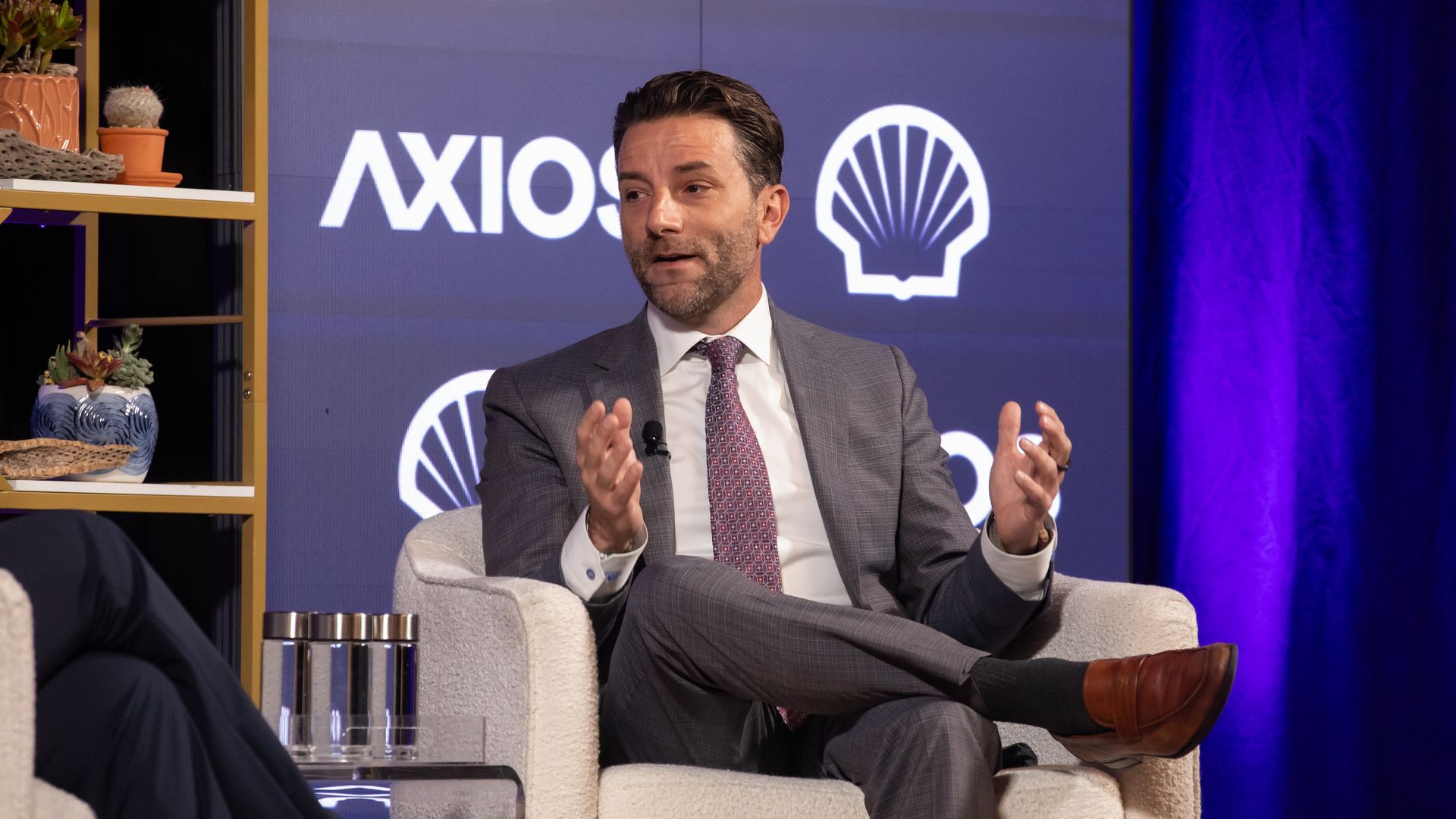 A man in a gray suit and maroon tie sits in a beige armchair on a stage with a blue "AXIOS" backdrop and logos; left shelves hold potted plants.