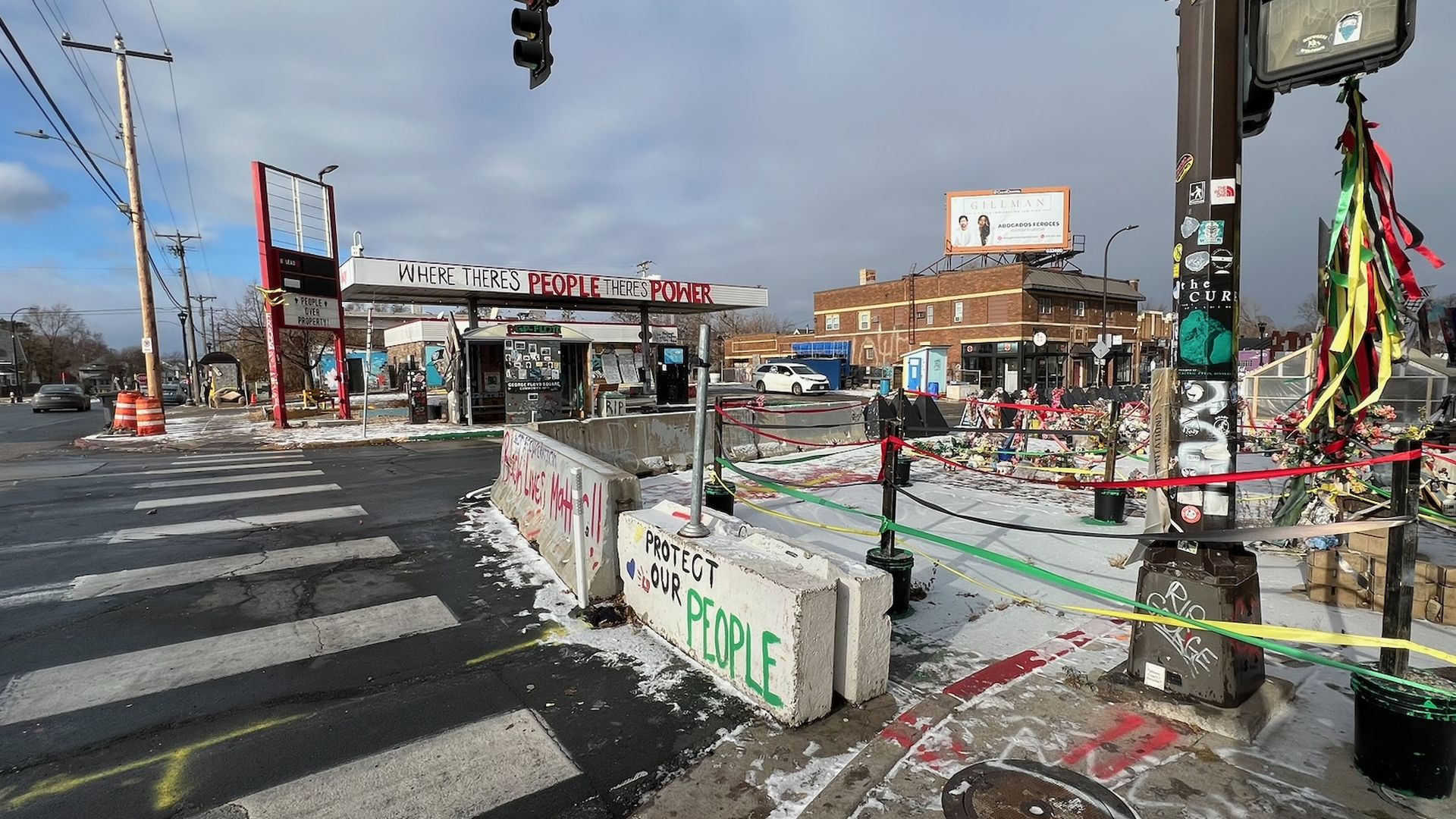 Urban intersection with a tall traffic light on the right, a crosswalk, and a memorial area with ribbons; a barrier reads "PROTECT OUR PEOPLE" and "WHERE THERE'S PEOPLE THERE'S POWER".