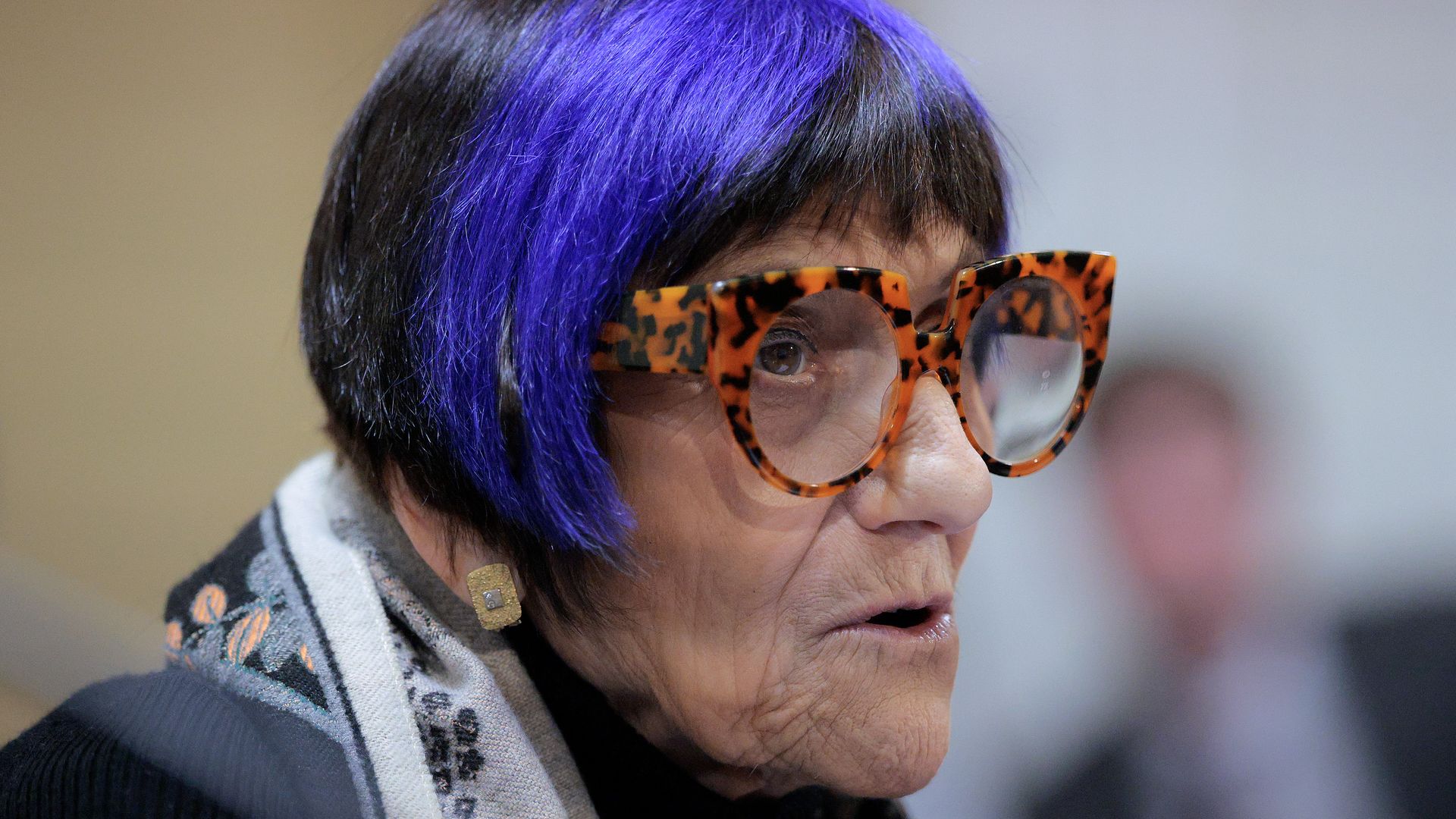 A woman with short black and purple hair, wearing large tortoiseshell glasses and a patterned scarf, speaking or in mid-conversation with a neutral background.