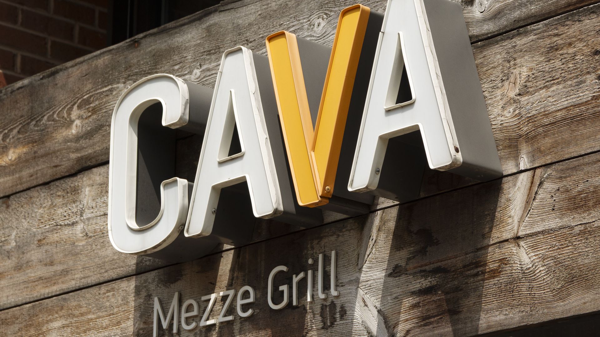 Close-up of a rustic wooden storefront sign with large white 3D letters and a bright yellow center piece, reading "CAVA Mezze Grill".
