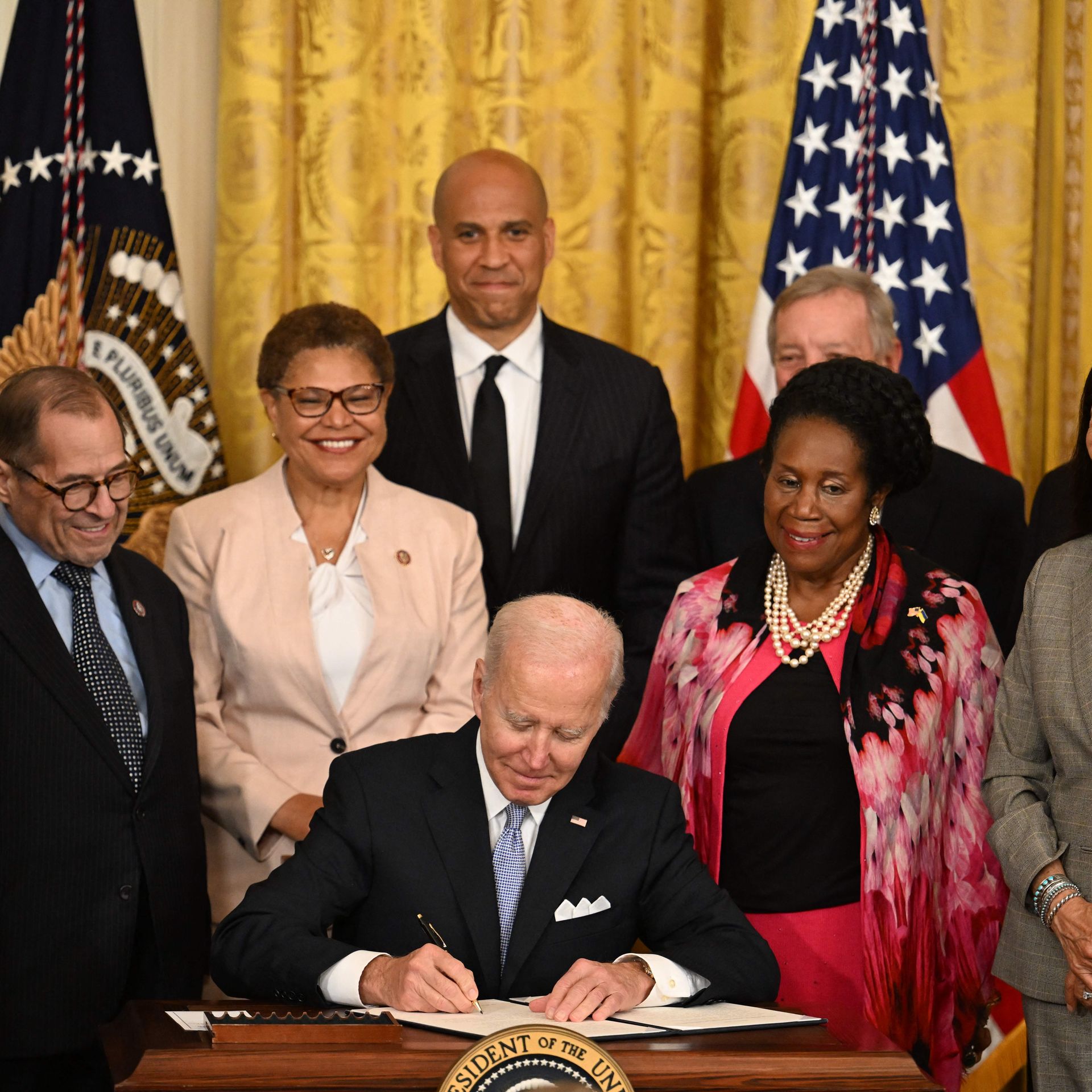 Photo of Joe Biden signing a bill at a desk while people stand around him smiling
