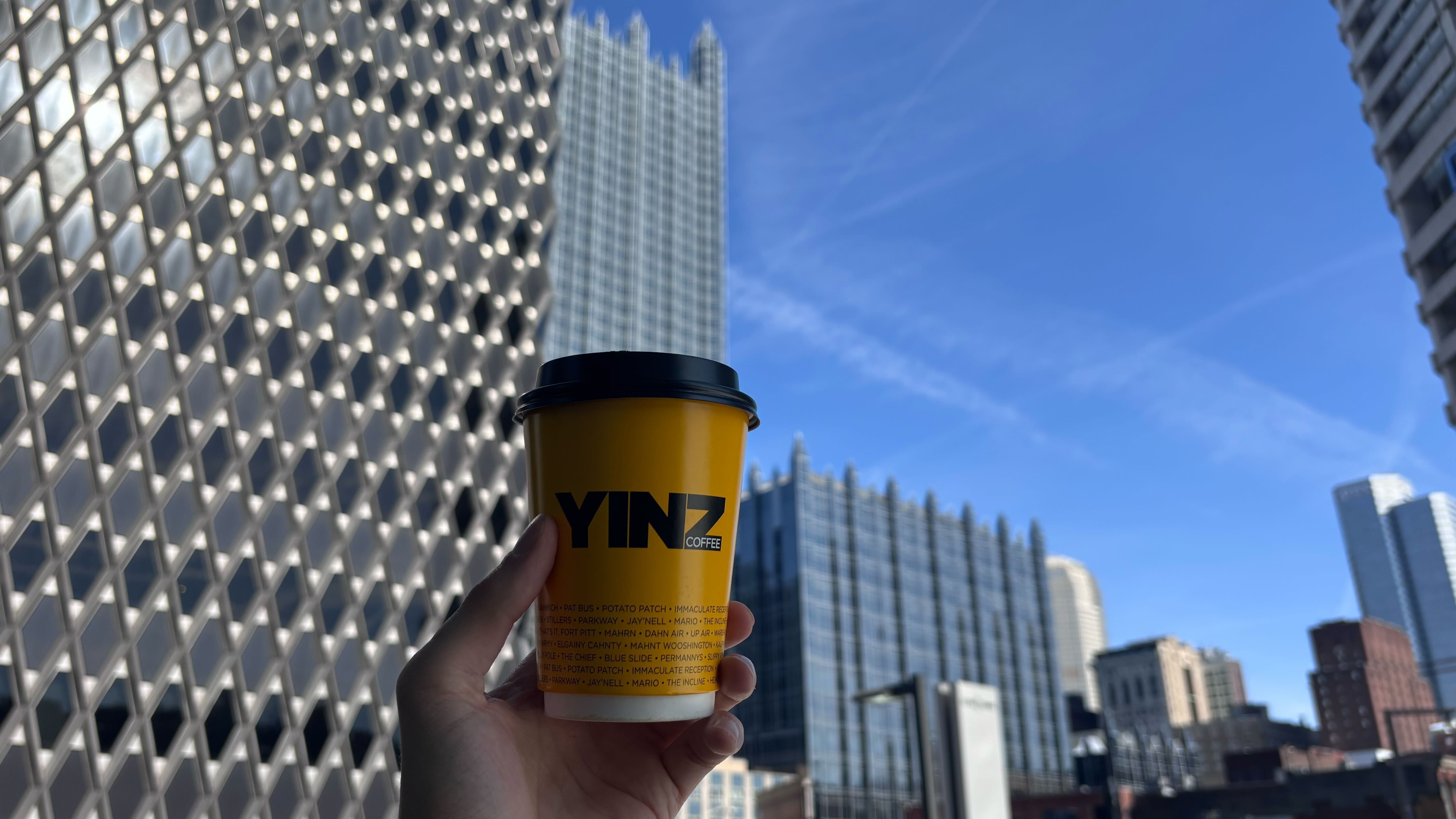 Hand holding a yellow coffee cup with black lid labeled "YINZ COFFEE" against a backdrop of modern city skyscrapers and a clear blue sky.
