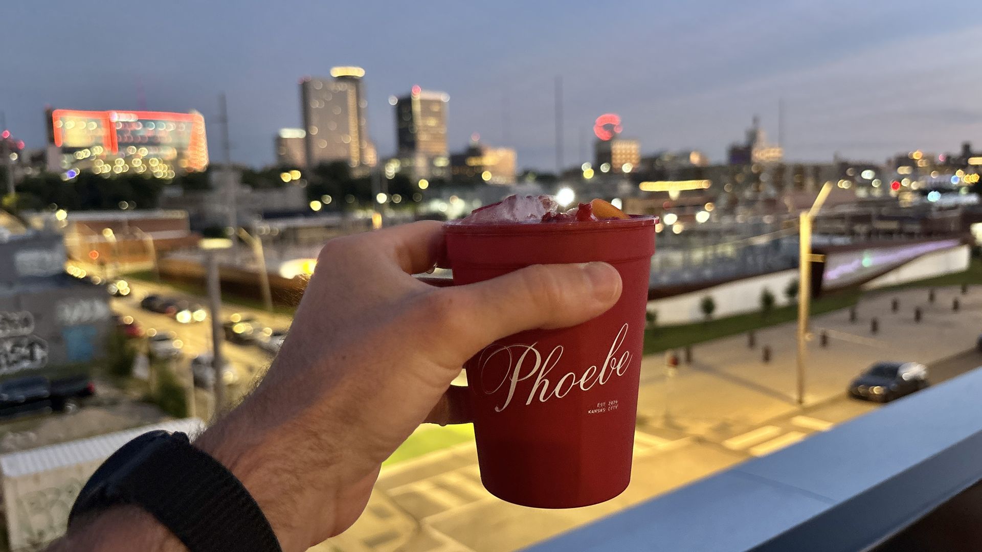 A hand in a dark sleeve holds a red cup with ice and fruit on a rooftop, while a city skyline blurs into colorful bokeh lights at dusk.