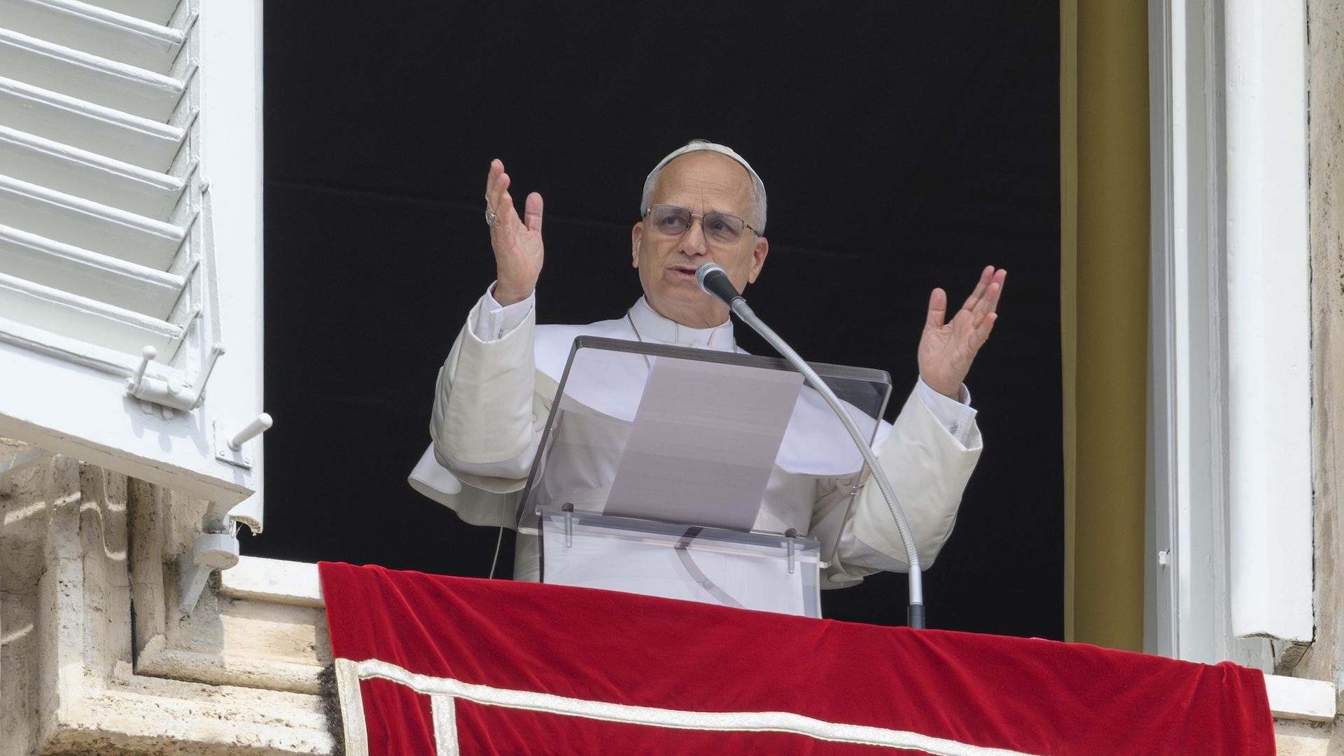 Pope Leo XIV at a lectern behind a microphone with a red drapery.