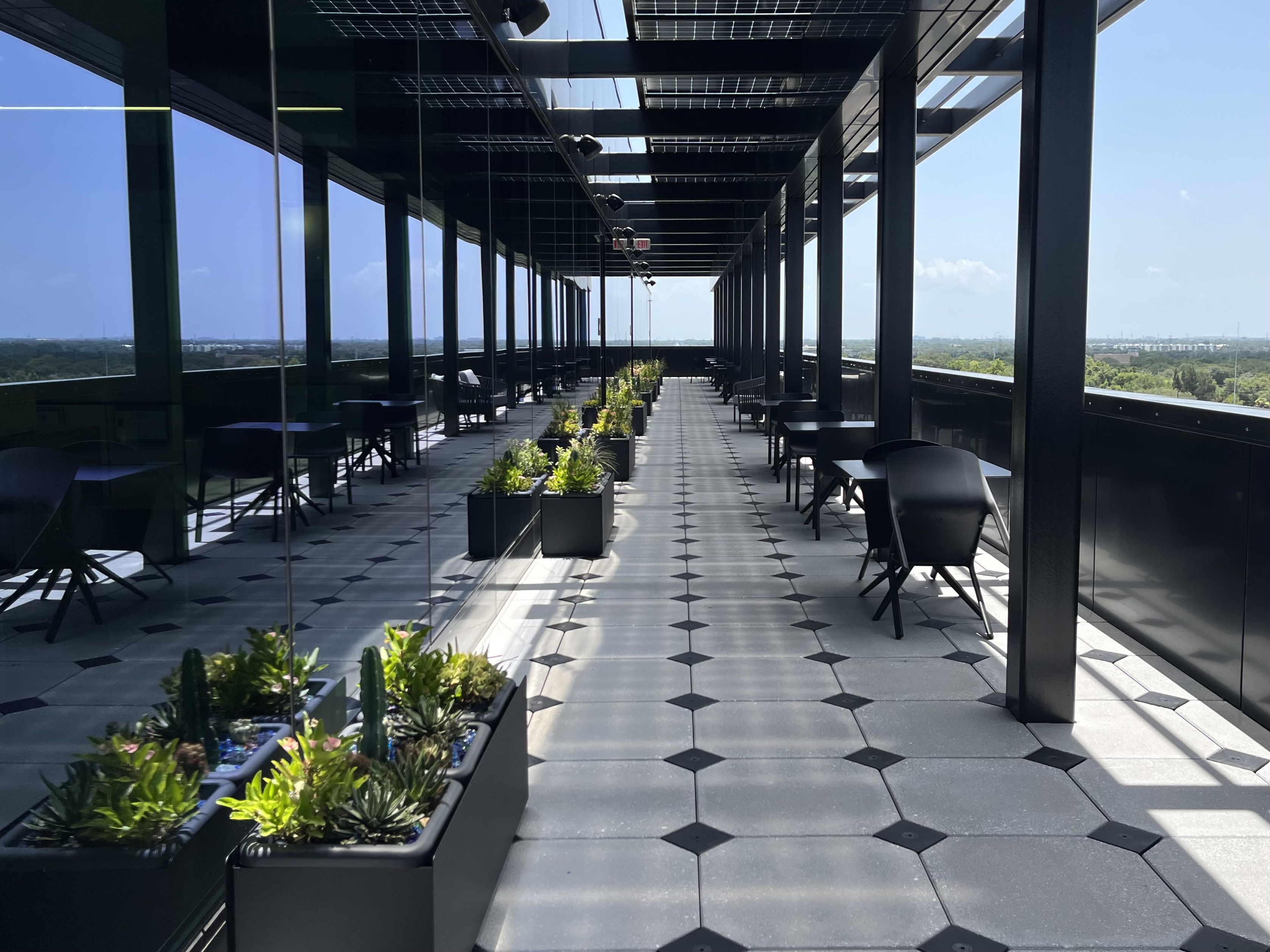 A rooftop terrace with glass walls and dark metal beams, rows of black tables and chairs, and long planters of green plants along a tiled floor; distant blue sky and cityscape beyond.