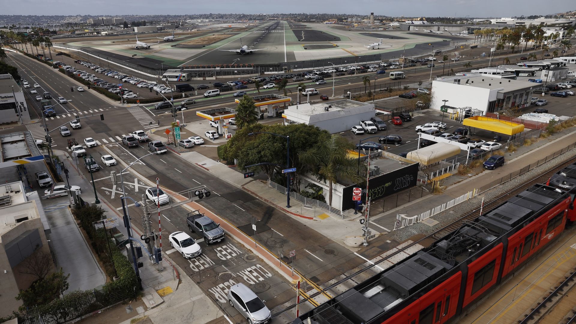 Airplanes line up waiting to depart from San Diego International Airport as traffic and a trolley travel pass by.