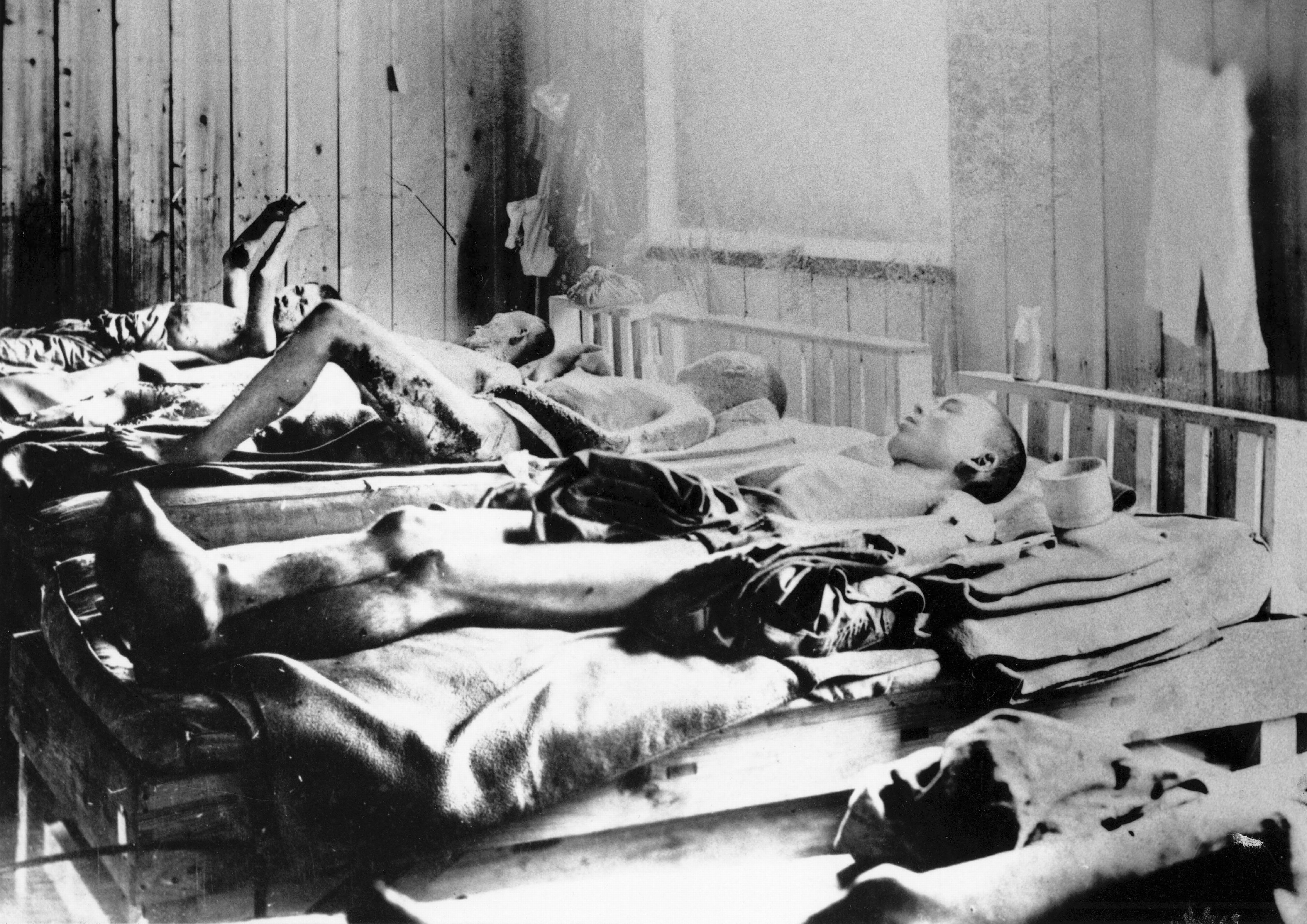 Black and white photo of emaciated men lying on beds in a wooden room, showing severe malnutrition or illness, with light coming from a window on the right side after the explosion of the atomic bomb at Hiroshima 1945.