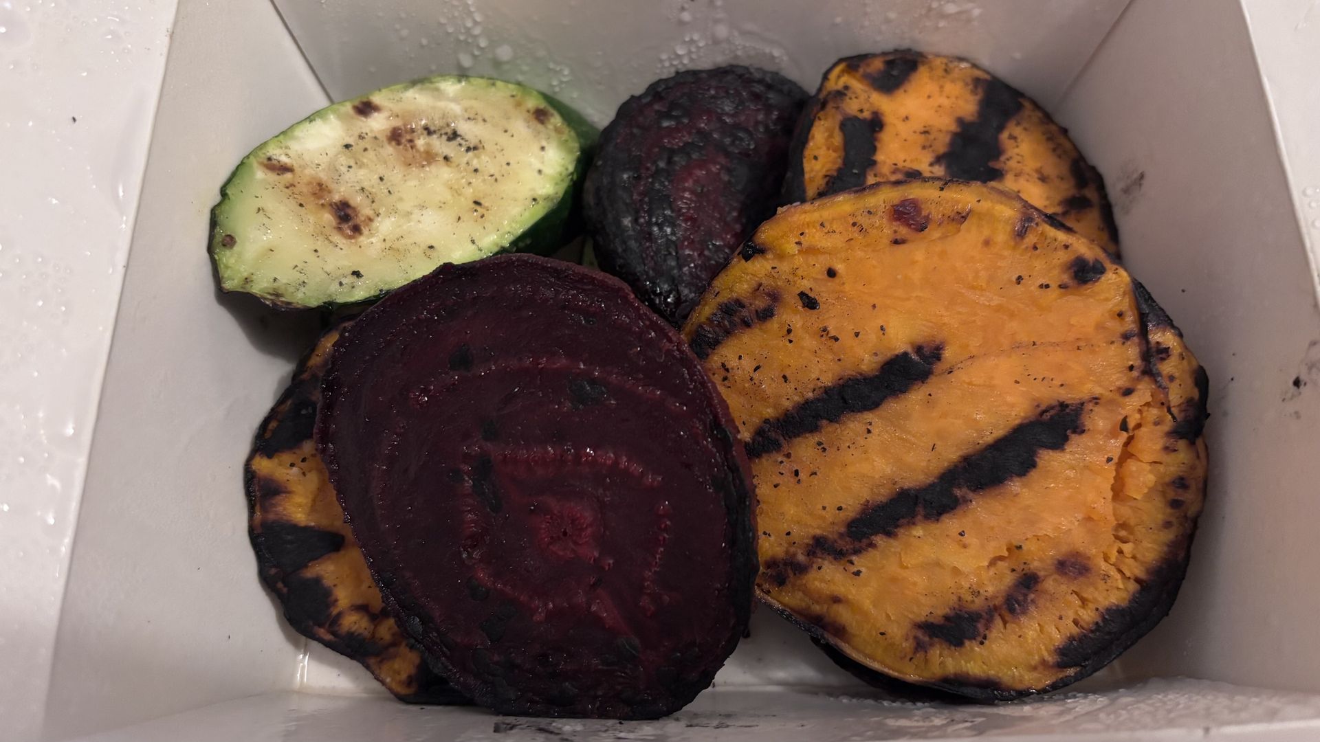 Grilled slices of sweet potato, beet, and zucchini with char marks served in a white container with droplets of moisture.