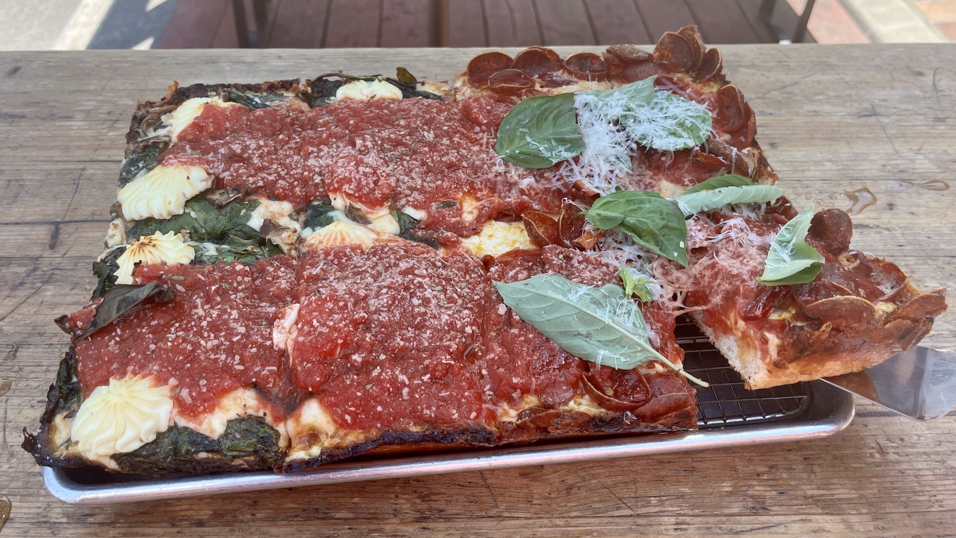 A detroit-style pizza with riccota, red sauce, pepperoni and fresh basil on a tray on a picnic table.