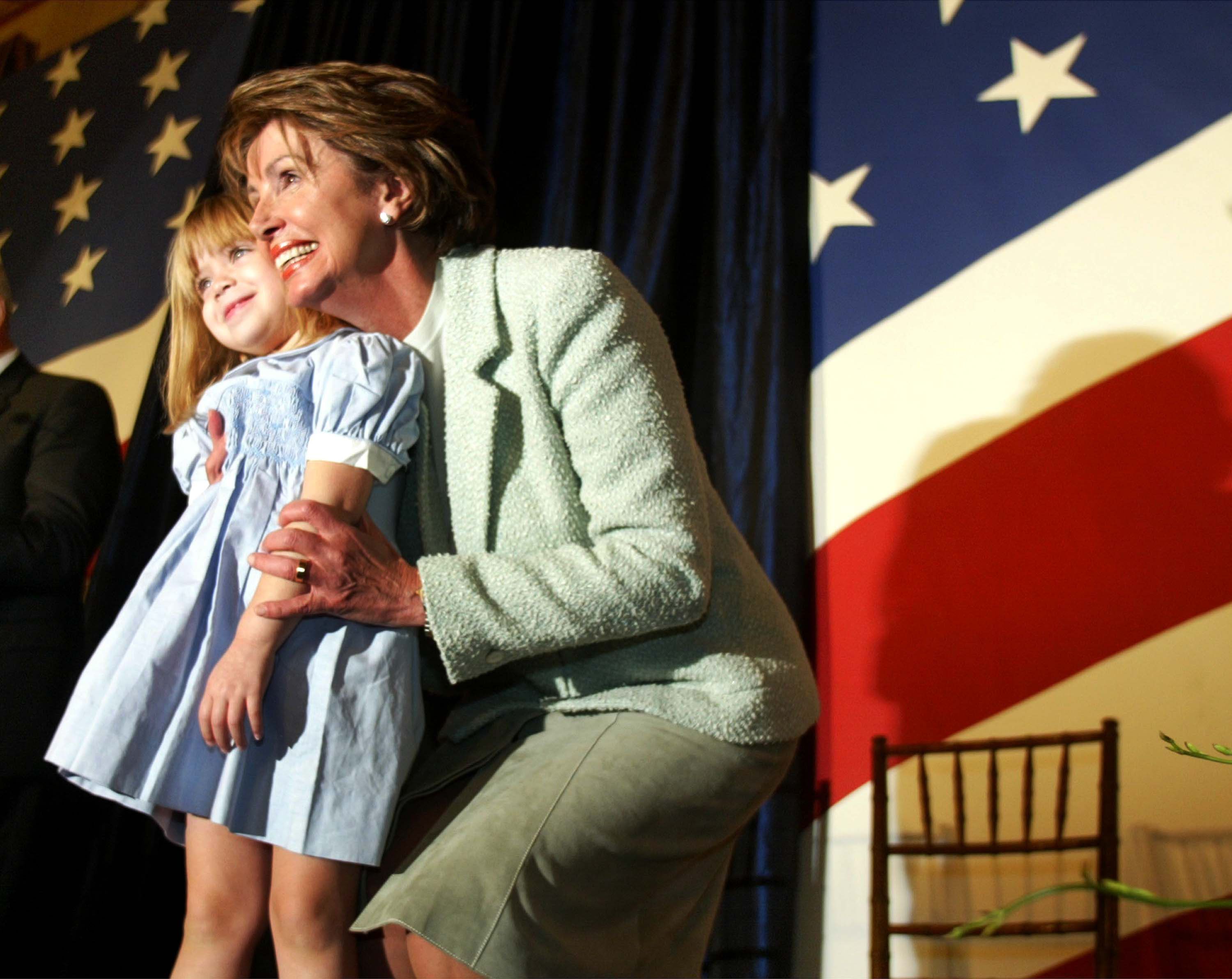 Nancy Pelosi embraces her young granddaughter during her 2003 swearing-in as House Democratic leader on Capitol Hill. An American flag print can be seen behind them.