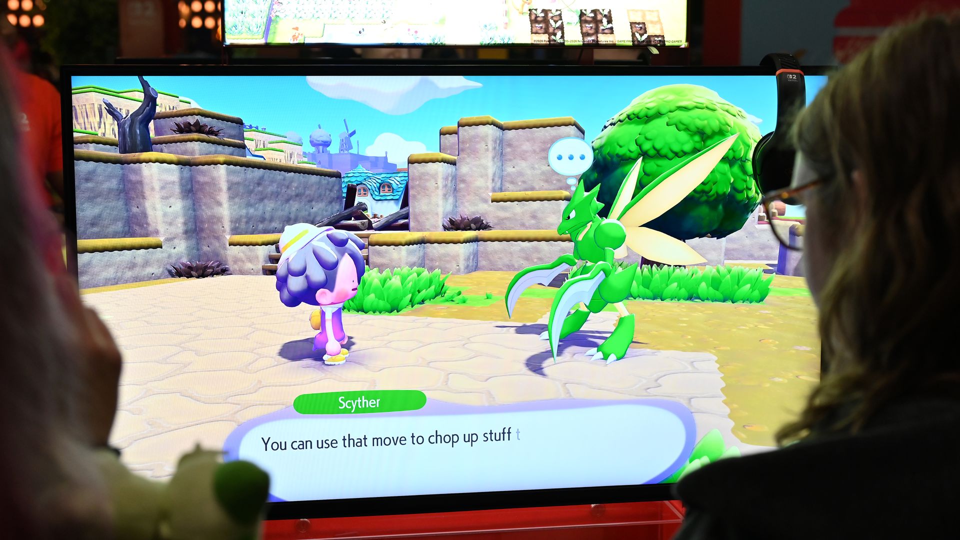 Two players watch a bright cartoon game on a large monitor: a green dragon-like creature faces a small purple-hatted character in a sunny stone courtyard, with a blue speech bubble and game text.