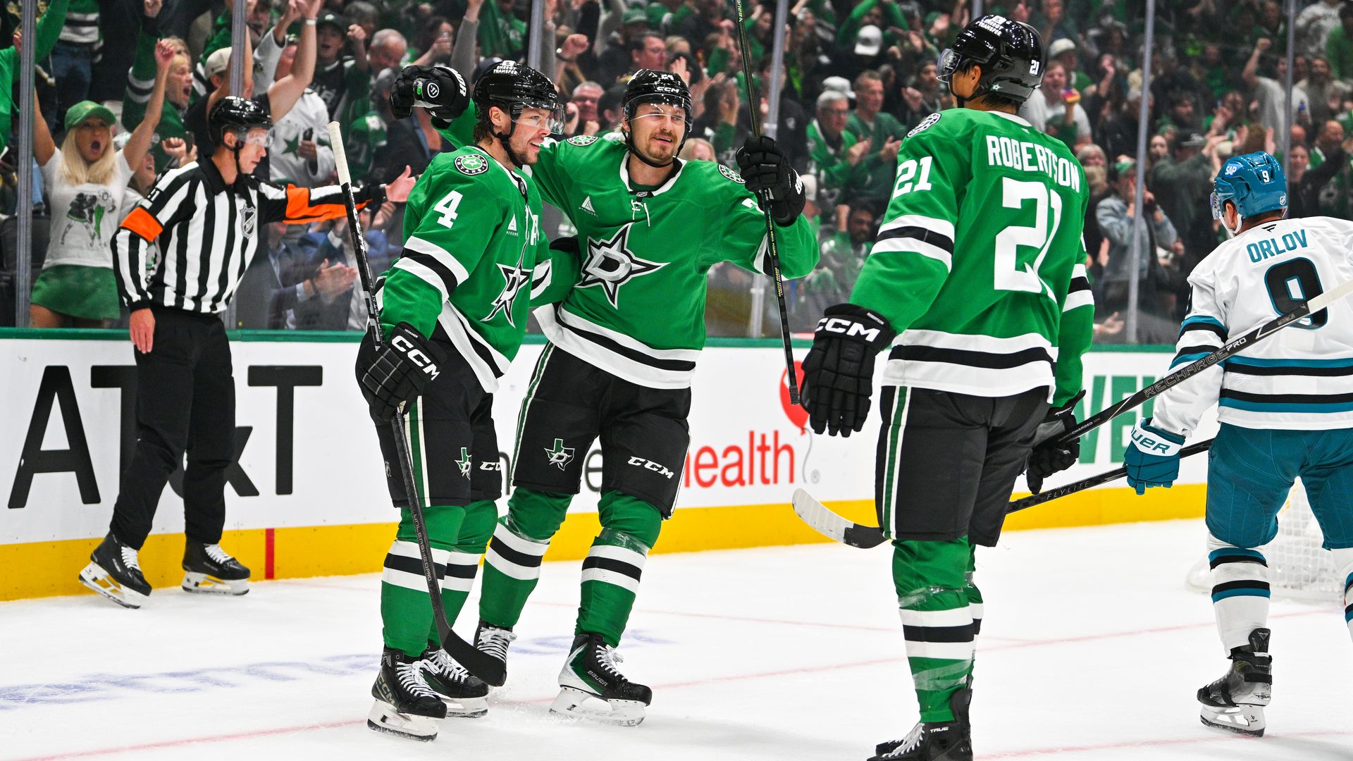 Dallas Stars players in green celebrate a goal on the ice; numbers 4 and 21 (Robertson) visible. Opponent Orlov in white and blue watches as the crowd cheers and a referee looks on.