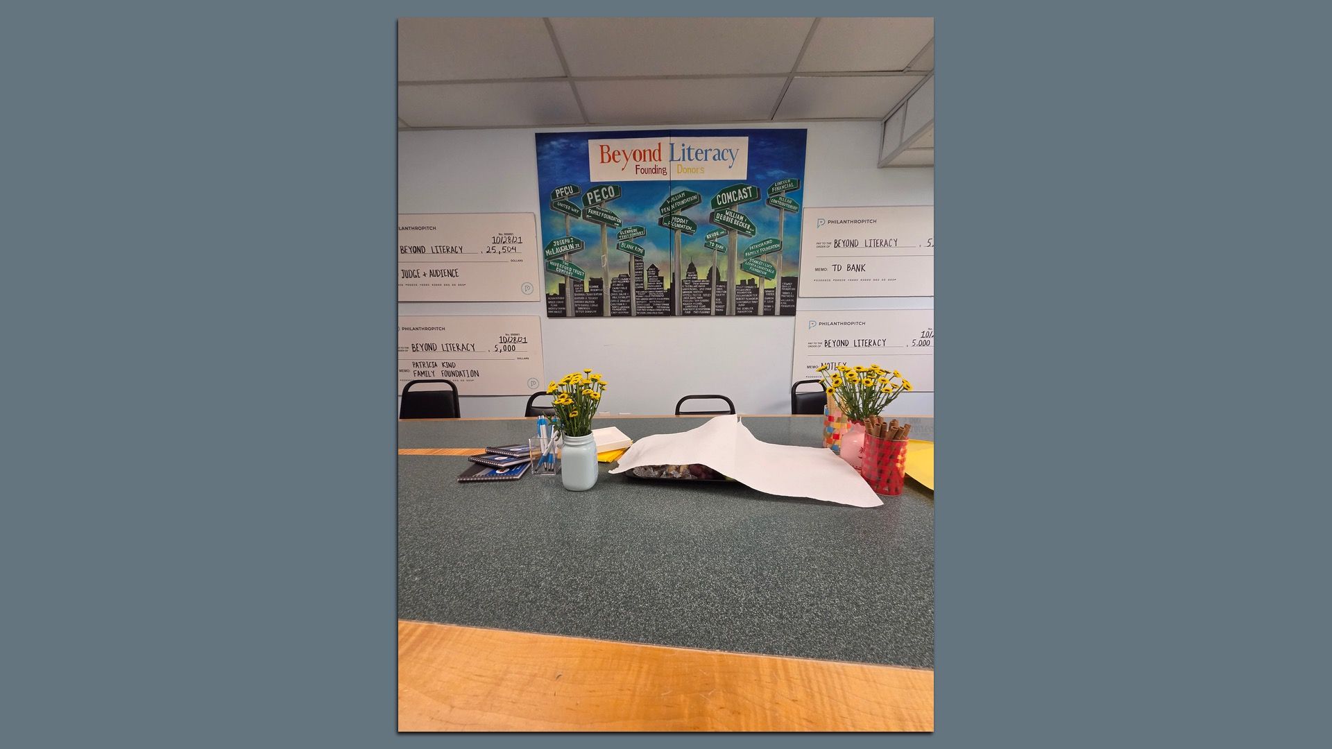 Reception desk with a large poster that reads "Beyond Literacy" and shows donor logos. A gray speckled counter holds yellow daisies, a mug of pens, scattered papers, and donation checks on the wall.