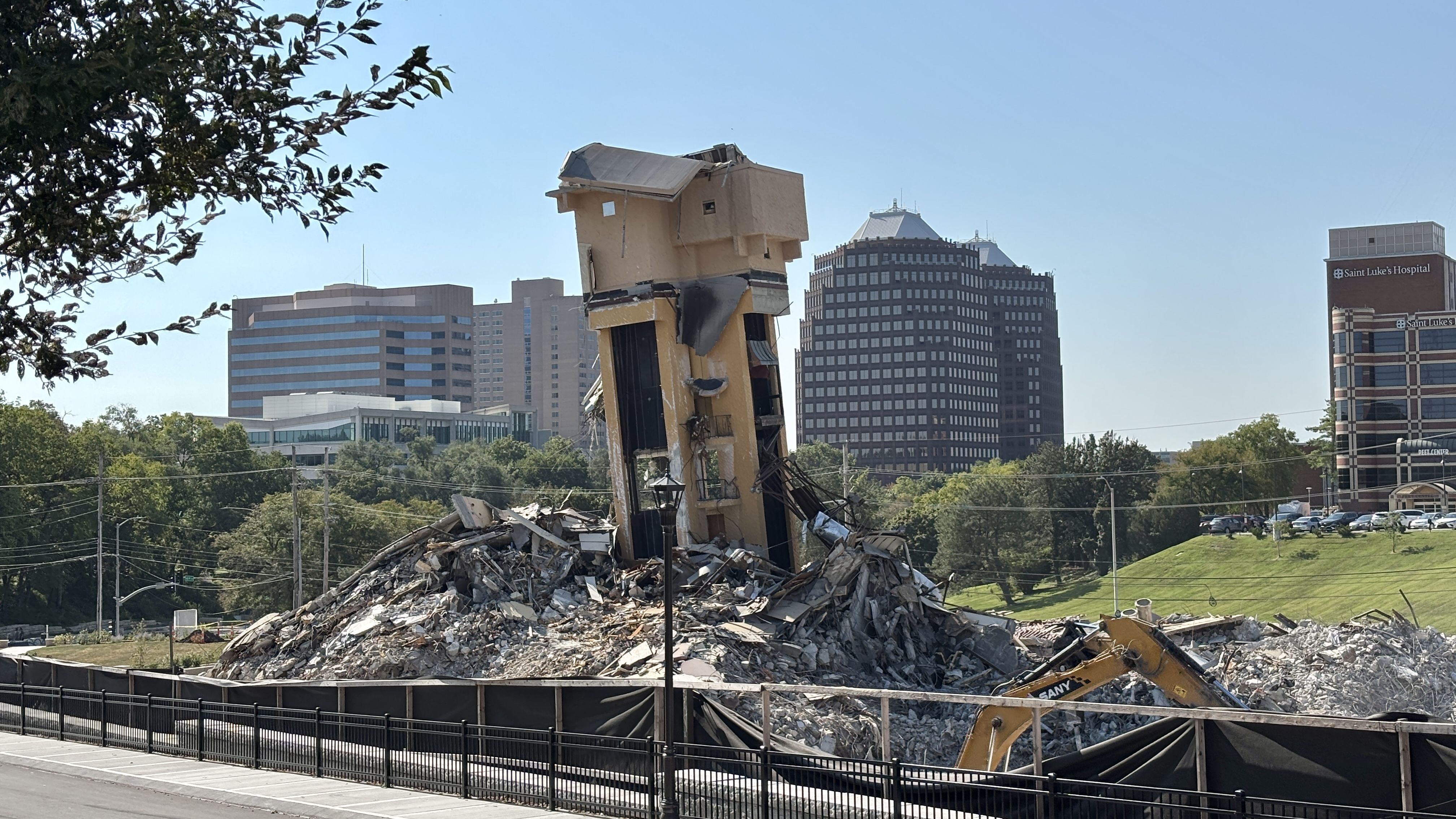 Beige building toppled into rubble at a demolition site; a yellow excavator arm visible. In the background are modern office towers and Saint Luke's Hospital; tree branches frame the left.