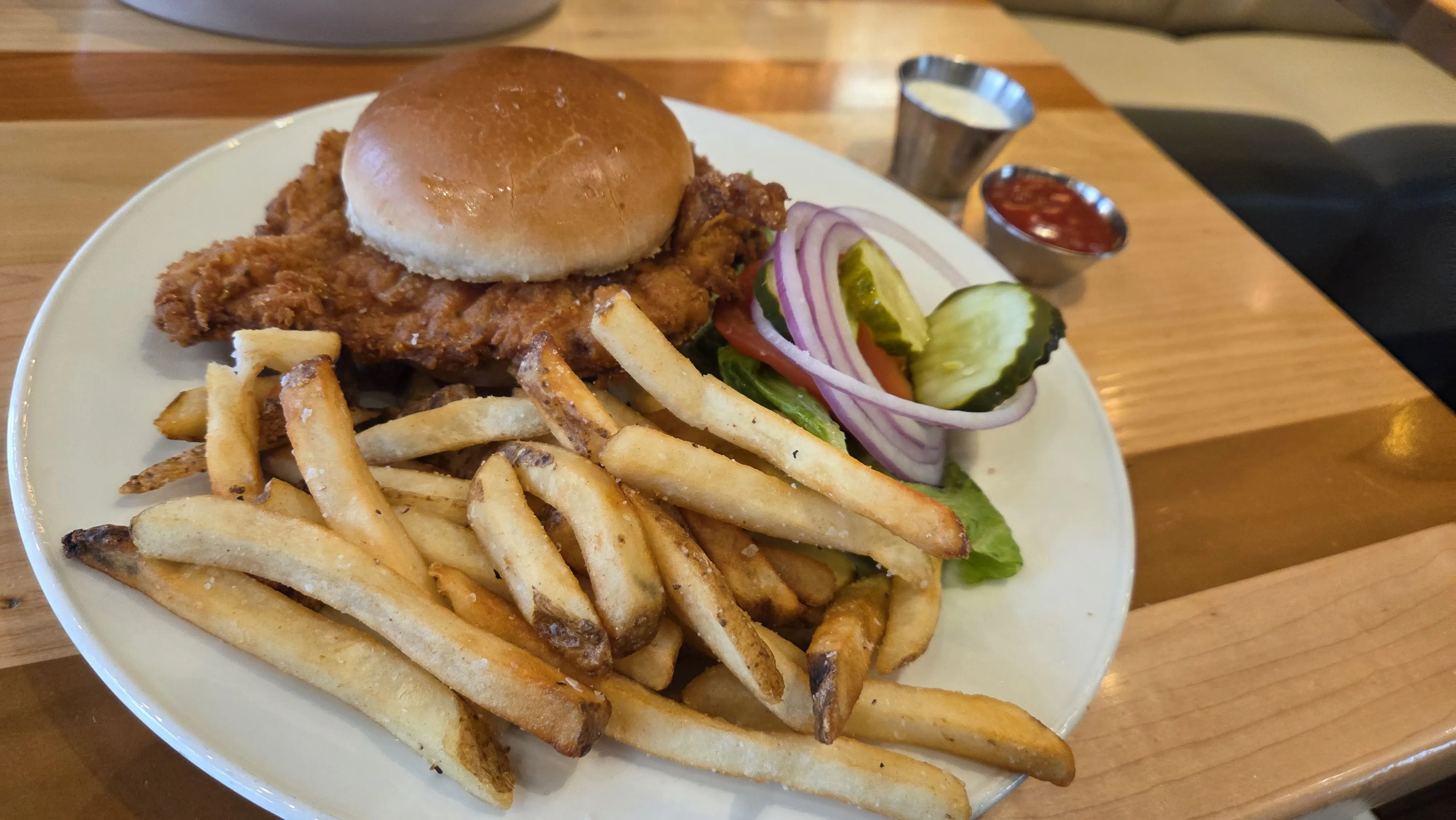 Fried chicken sandwich with lettuce, tomato, red onion, pickle, and a side of seasoned French fries on a white plate, accompanied by ketchup and ranch dipping sauces.