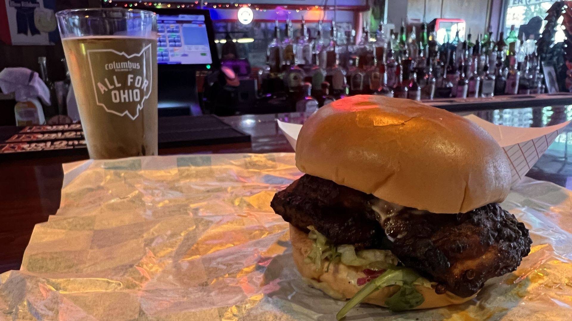 Large burger with a thick, dark patty and melted cheese, lettuce on a toasted bun sits on parchment at a bar. A beer glass labeled "Columbus All For Ohio" sits to the left; neon lights glow.