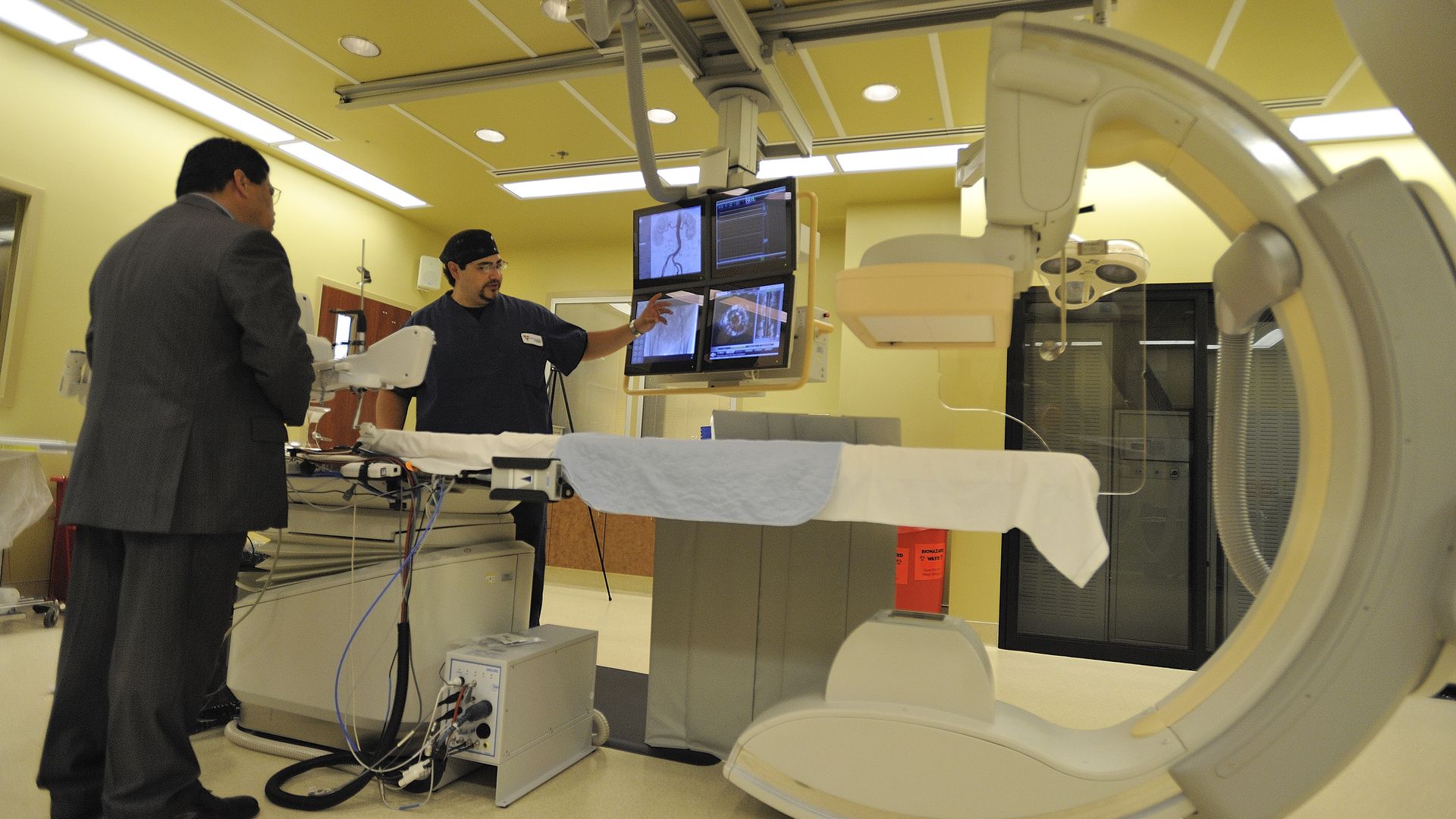 A health care technician points to computer screens near a cardiac cath machine.