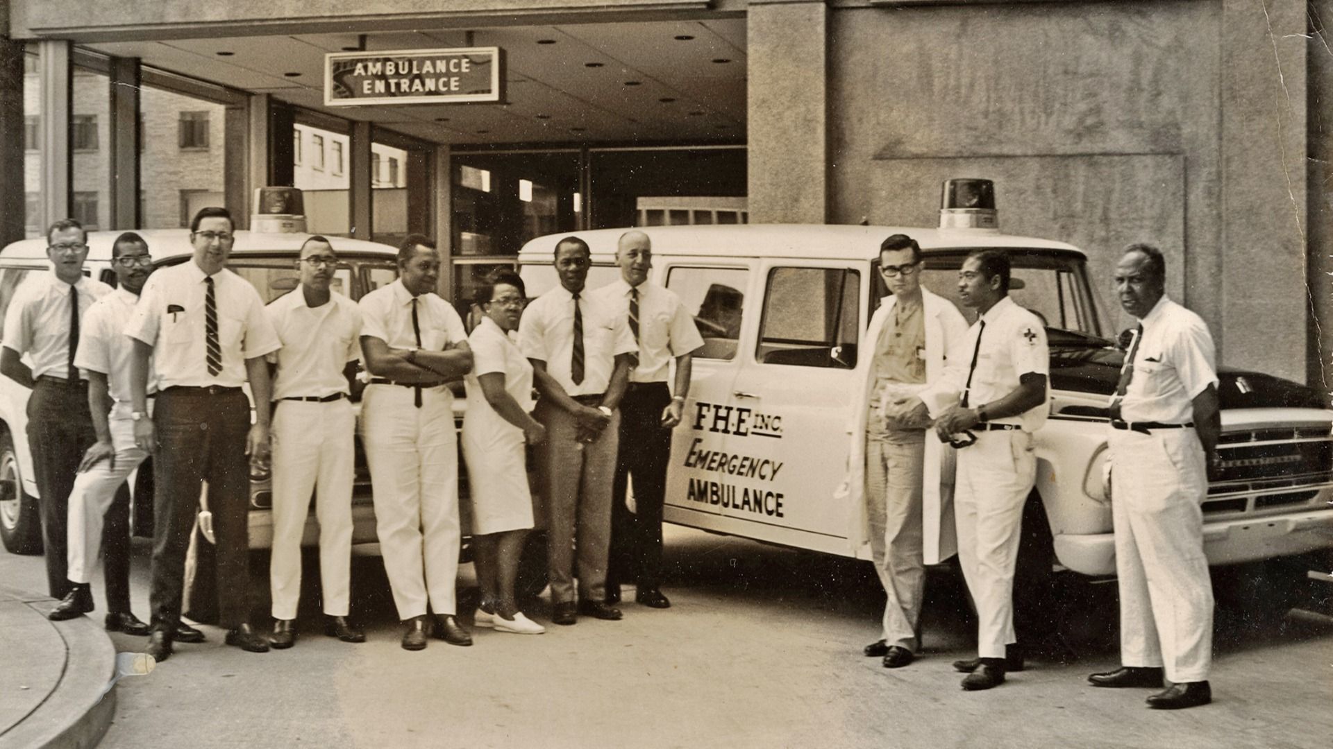 Black-and-white photo of thirteen men and one woman standing in front of two emergency ambulances under an "Ambulance Entrance" sign, all dressed in professional attire.