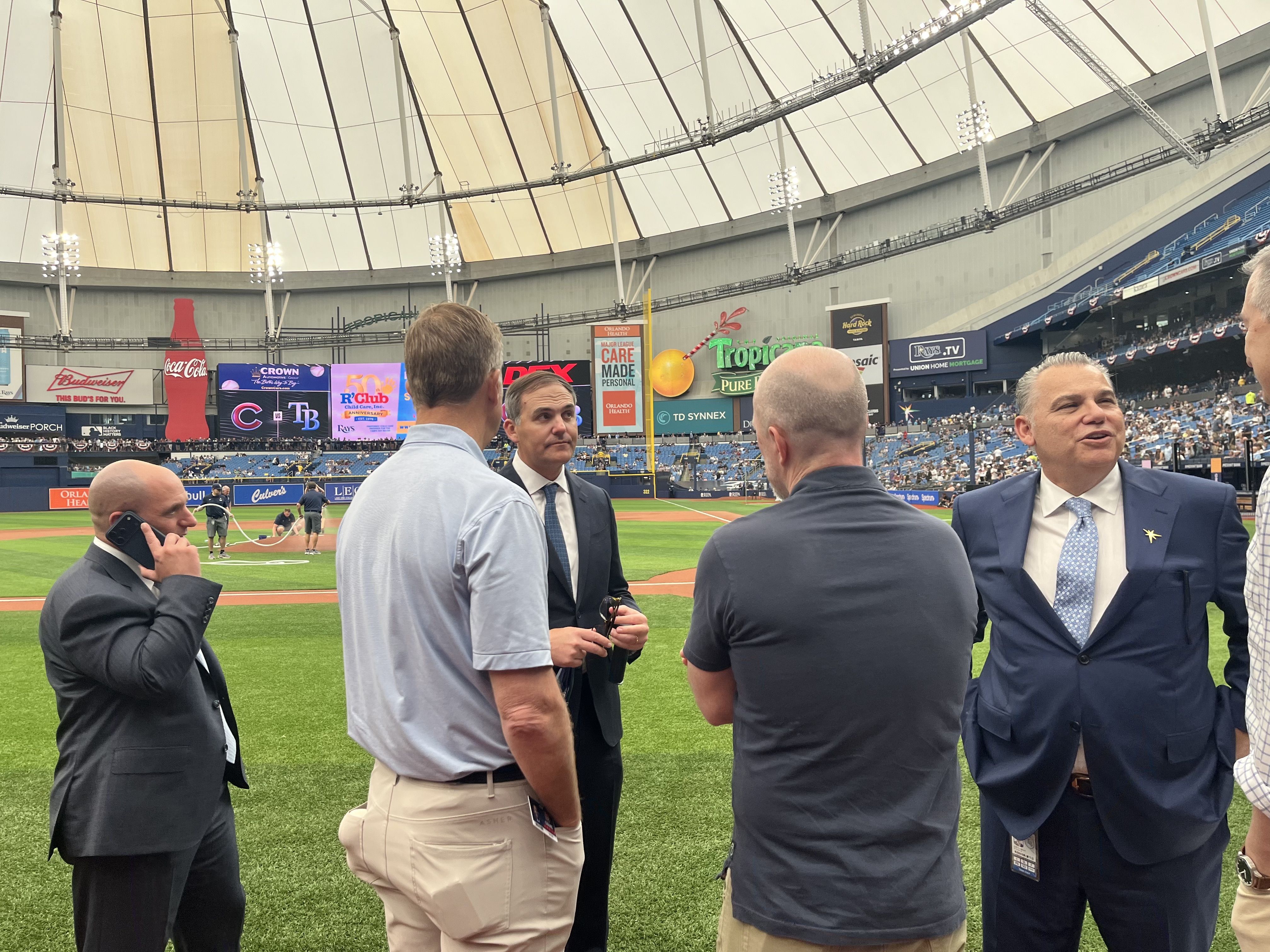 Inside a large domed baseball stadium, men in suits chat on the grass. One man on the left talks on a cellphone while others converse near sponsor banners.