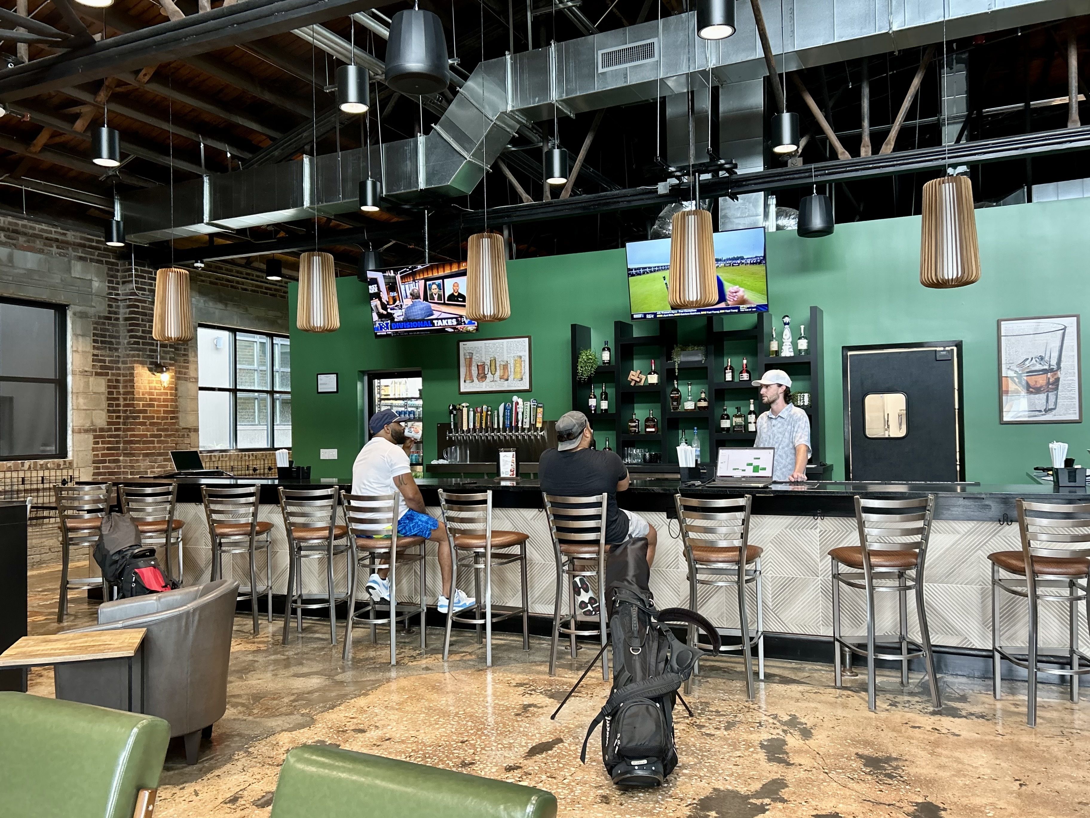 Two men sitting at a bar with a bartender serving; green wall with shelves of liquor, multiple pendant lights, TVs showing sports, and a golf bag in front of the bar in an industrial-style space.