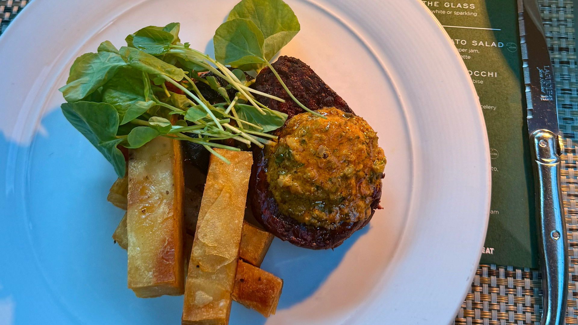 White plate with a vegan dish including greens, fried potato sticks, and a patty topped with sauce. Nearby is a cocktail with an orange slice, a lit candle, flowers, and a vegan menu.