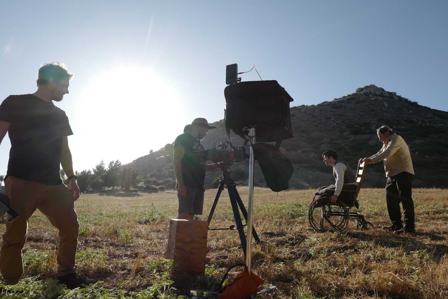Sunlit hillside film set with four crew members, one adjusting a tripod camera, another helping a man in a wheelchair, and a large light on a stand nearby.