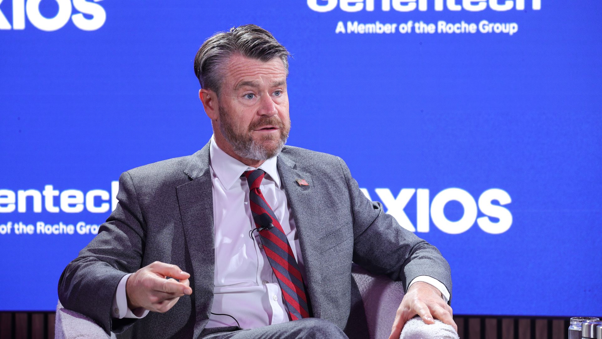 A man in a gray suit with a red striped tie sits on a stage, in front of a bright blue backdrop that reads Genentech and Axios.