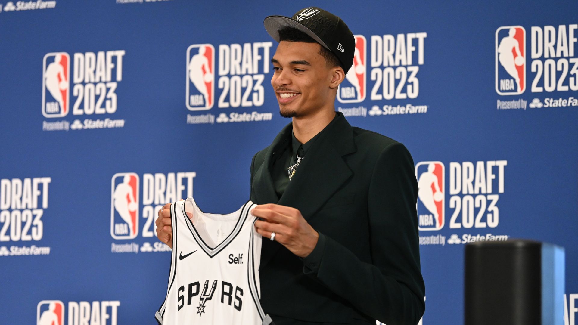 Wemby holds up his No. 1 Spurs jersey in white in front of an NBA Draft 2023 backdrop.