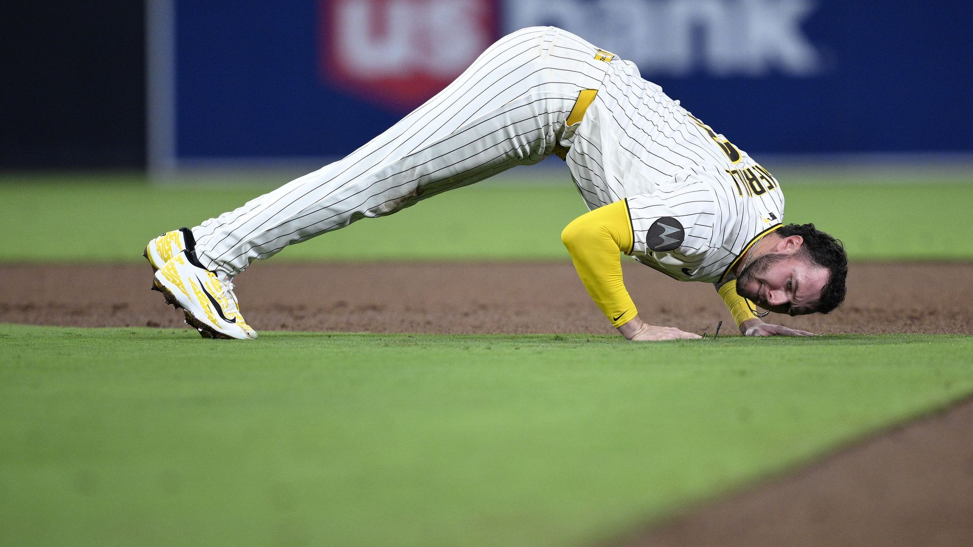 San Diego Padres player Jackson Merrill in a white pinstriped uniform with yellow accents dives onto the infield dirt, hands touching the ground, with green grass and a blue outfield wall in the background.