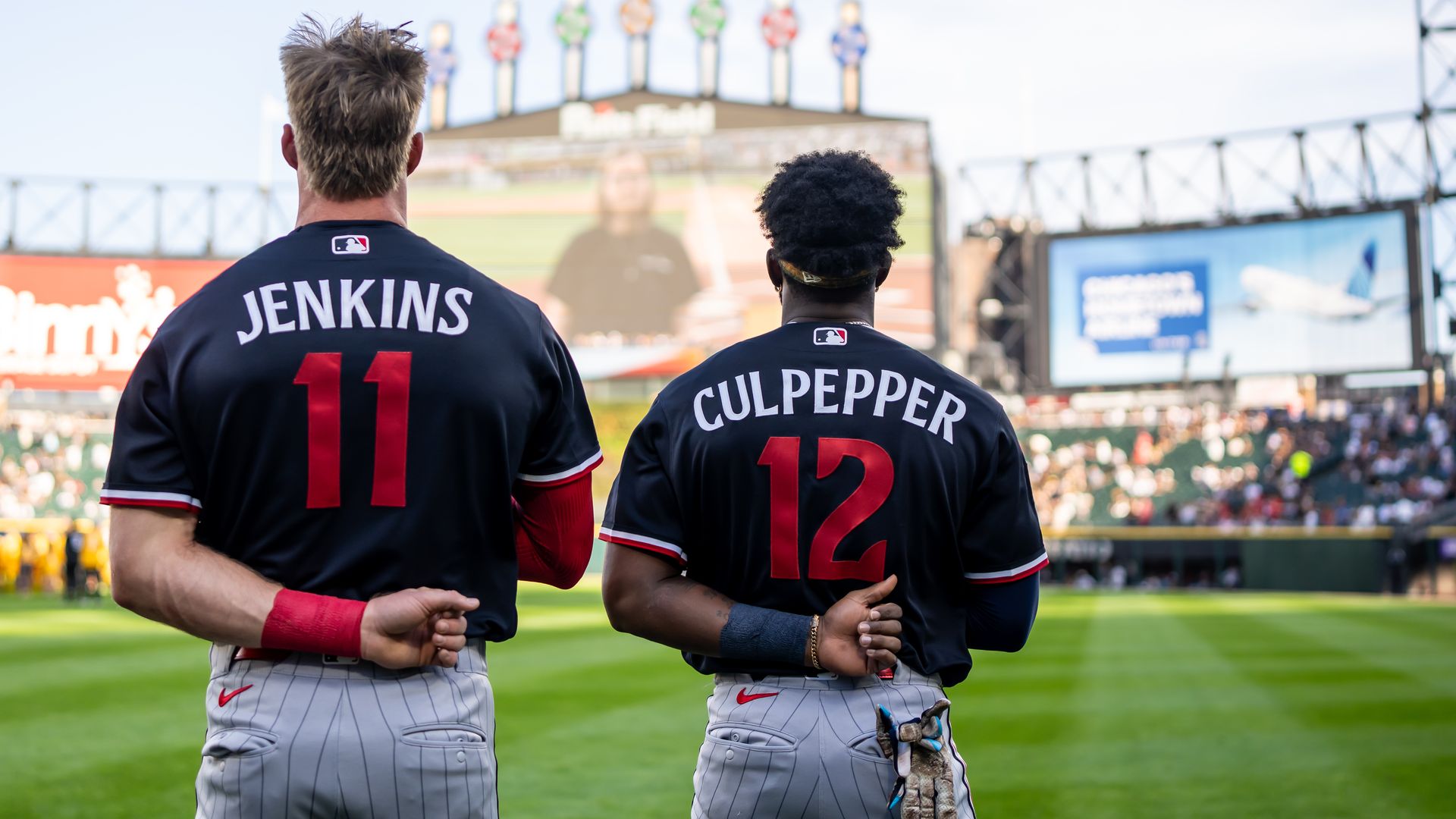 Two baseball players seen from behind on a sunny field, navy jerseys reading JENKINS and CULPEPPER with numbers 11 and 12; a blurred scoreboard and crowd in the background.