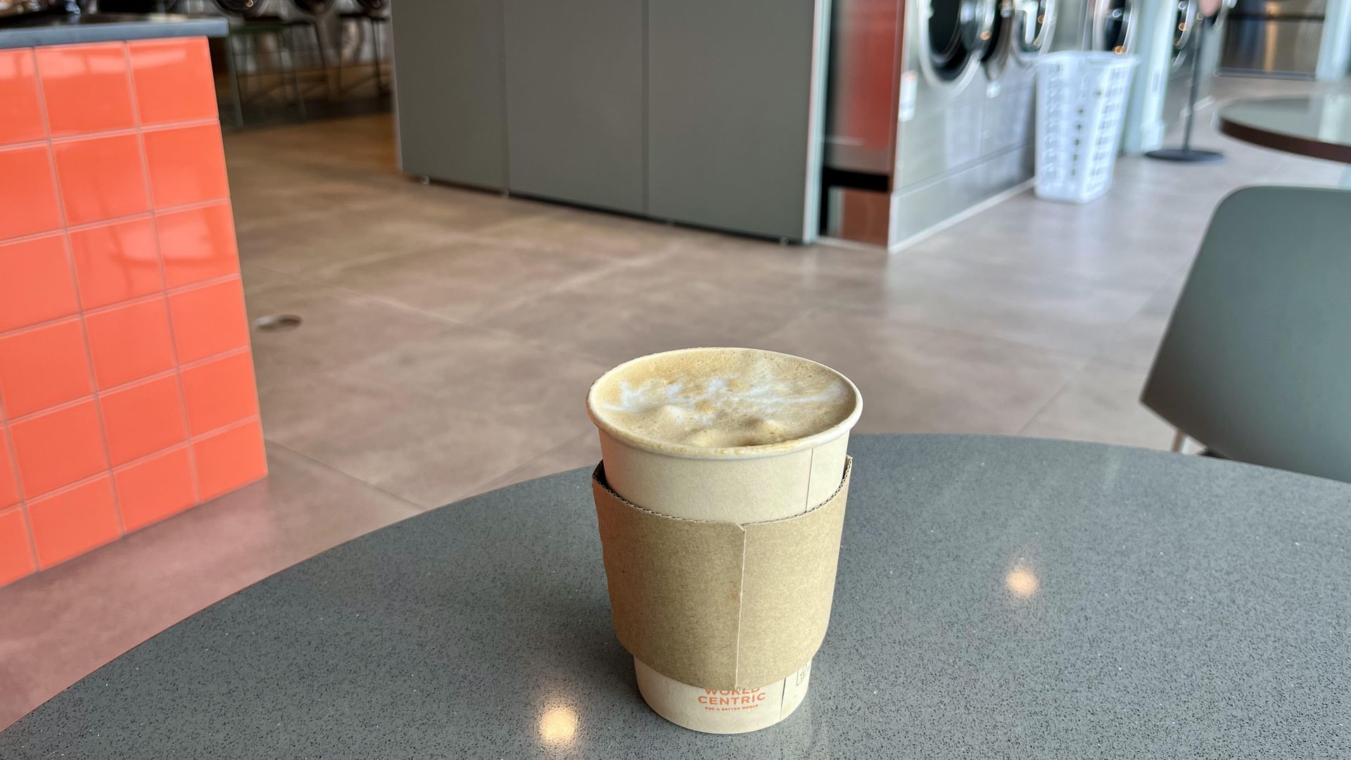 A takeaway coffee cup with a cardboard sleeve sits on a speckled gray table inside Soap Laundry Lounge in San Antonio, with front-loading washing machines and a red tile wall in the background.