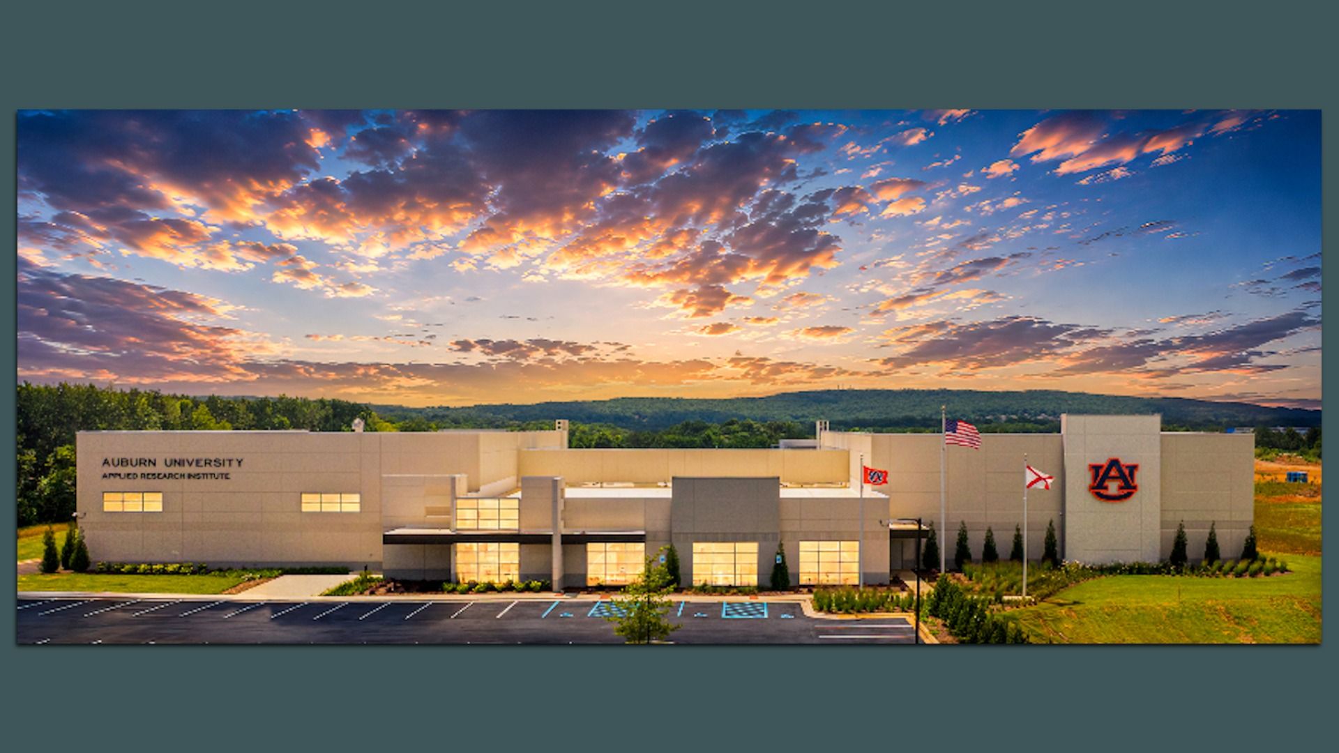 Wide view of the Auburn University Applied Research Institute building at sunset; beige façade with the AU logo and flagpoles, a parking lot in front, and a dramatic orange-pink sky with purple clouds.