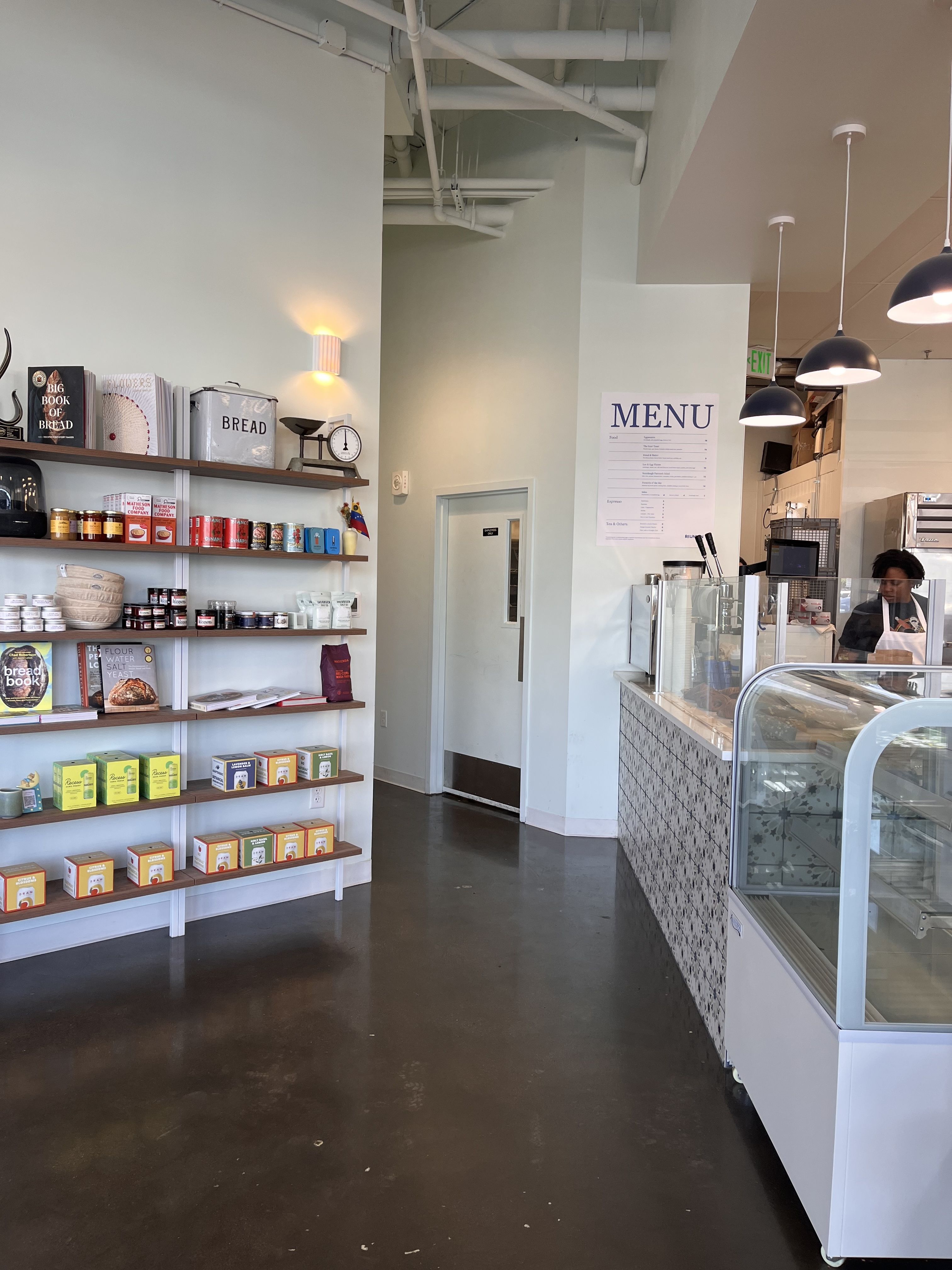 Bright cafe interior with pale mint walls and exposed white pipes. Left shelves hold colorful boxes and jars; right features a glass display counter and a staff member at the counter, with a MENU sign on the wall.