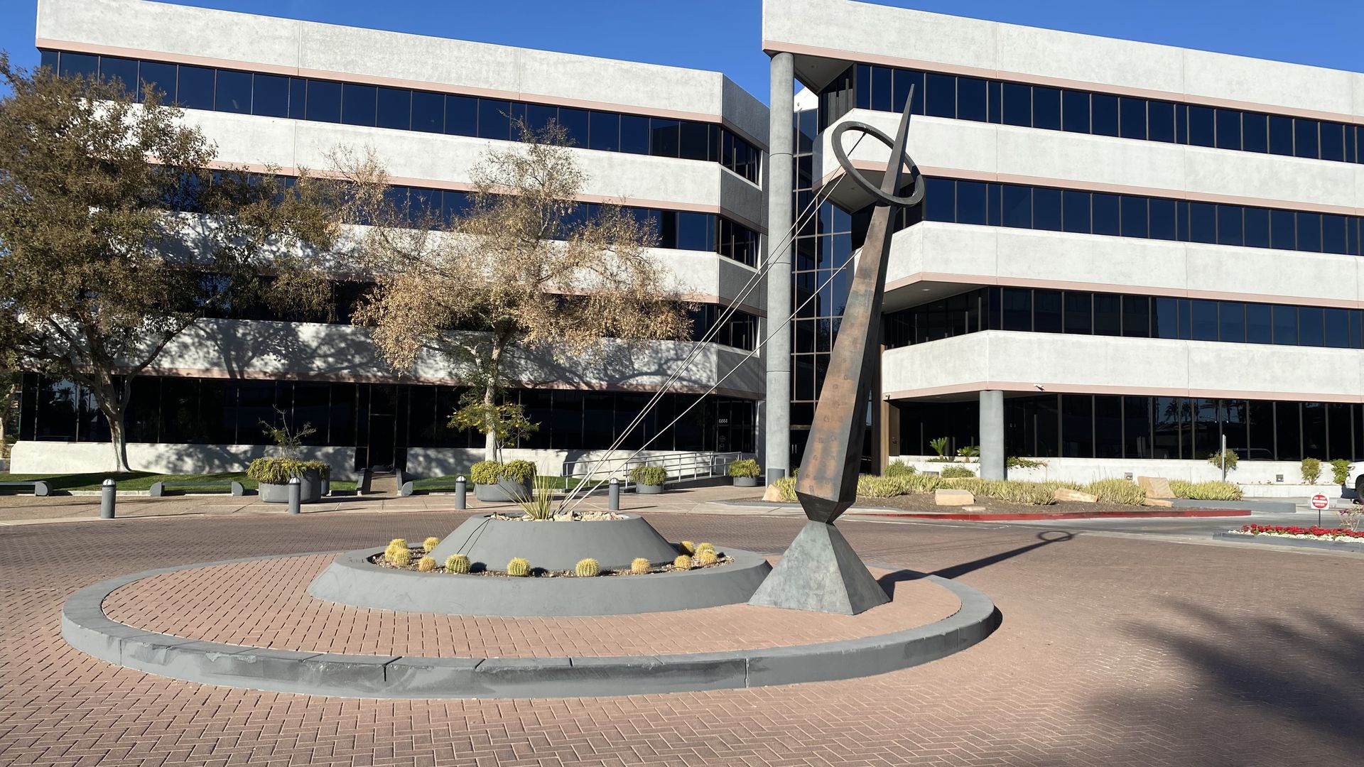A sculpture consisting of a leaning obelisk with a ring over the top that's held in place by three cables that are anchored to a circular pedestal on the ground. 
