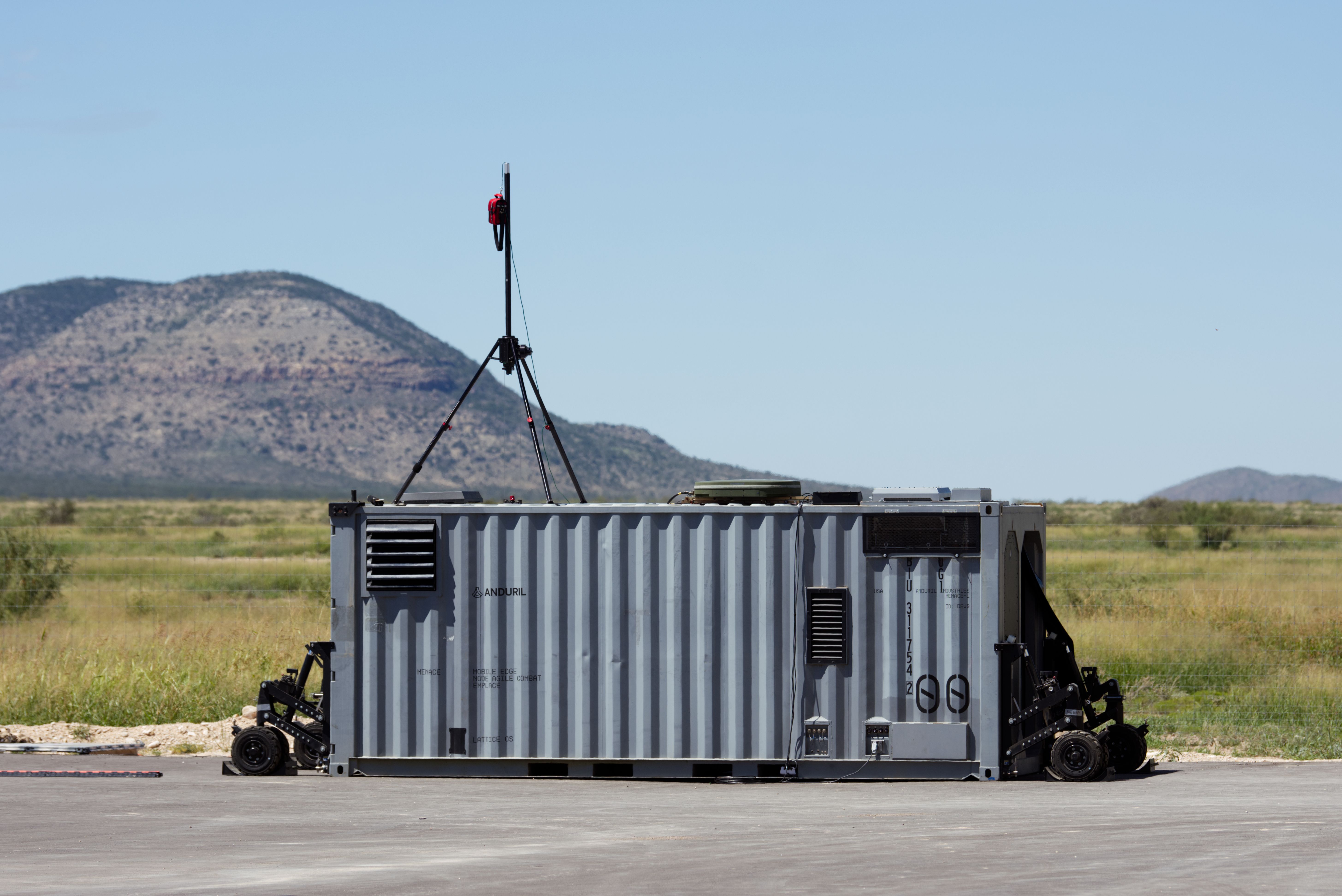 An upfitted shipping container sits on a runway. The sky behind it is blue. A bluff is seen to the back left. Grass is in the middle ground.