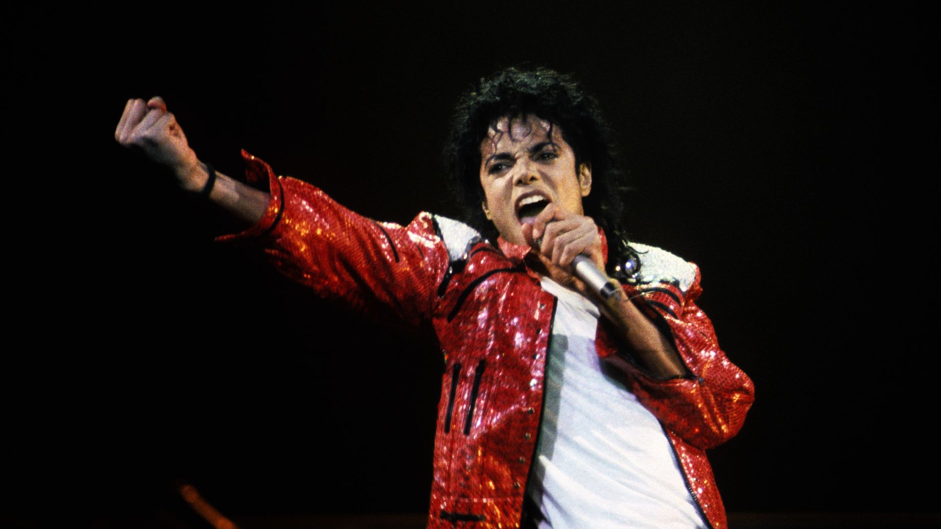 Male singer on a dark stage wearing a bright red sequined jacket and white shirt, singing into a microphone with his arm raised.