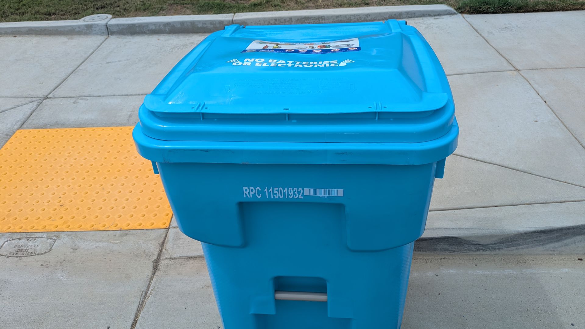 Blue recycling bin on a concrete sidewalk beside a yellow tactile paving strip; grass and curb behind it. The lid shows a sticker and reads "NO BATTERIES OR ELECTRONICS".
