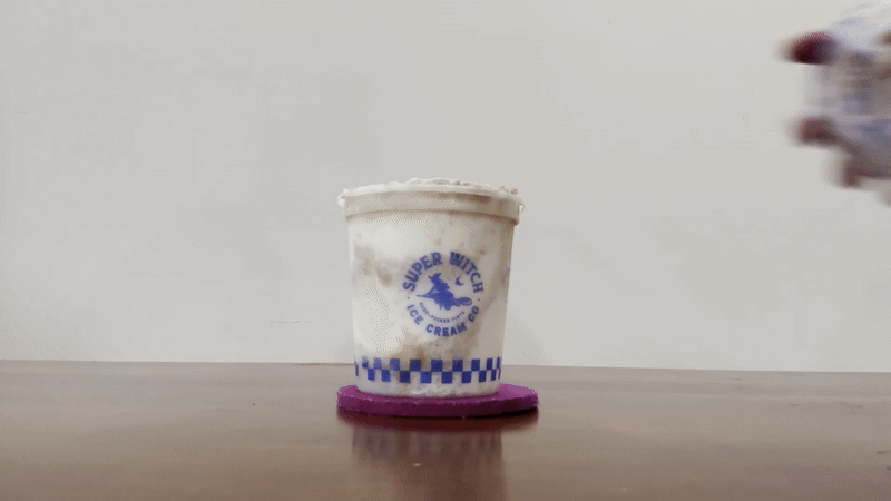Container of Super Witch Ice Cream Co. with white and brown swirled ice cream on a purple coaster against a plain white background.