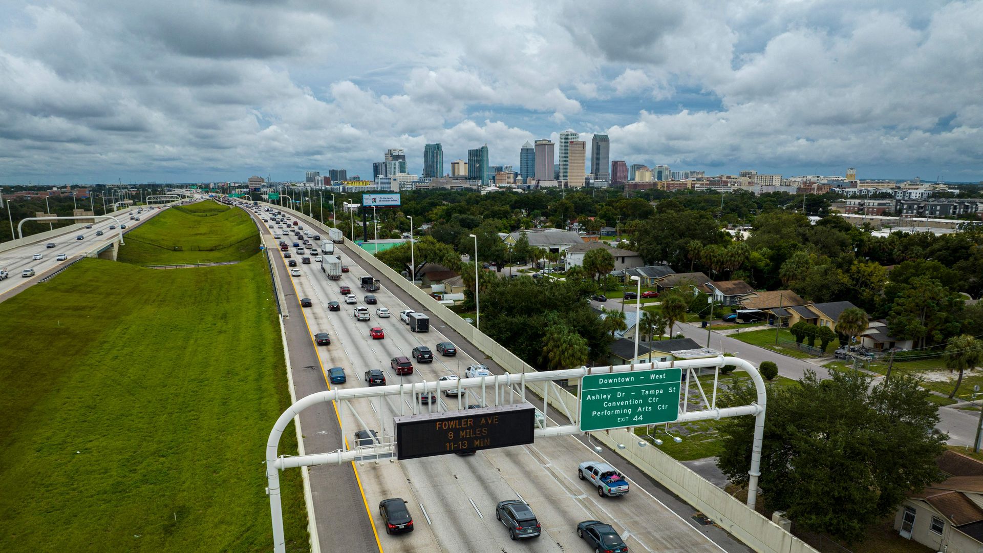 Traffic on a multi-lane highway leading into a city skyline with tall buildings under a cloudy sky, green grass median, and residential area with trees on the right.
