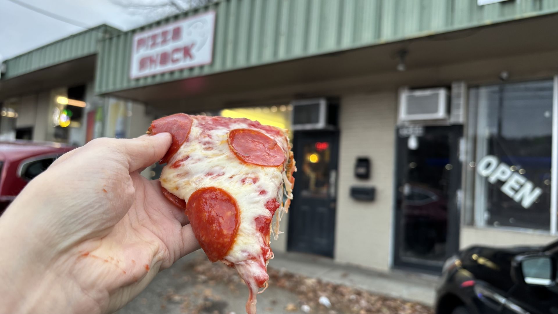 Hand holding a pepperoni pizza slice with melted cheese in front of a green building with a blurred 